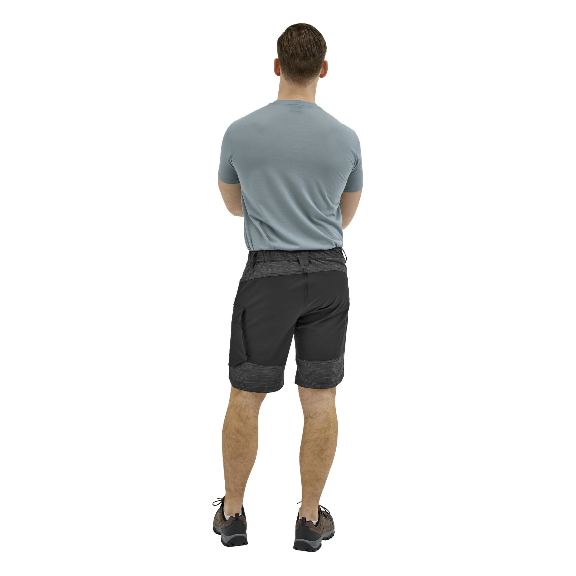 En man står med ryggen mot kameran, armarna löst korsade bakom sig, iklädd en ljusblå t-shirt, svarta shorts och sneakers.