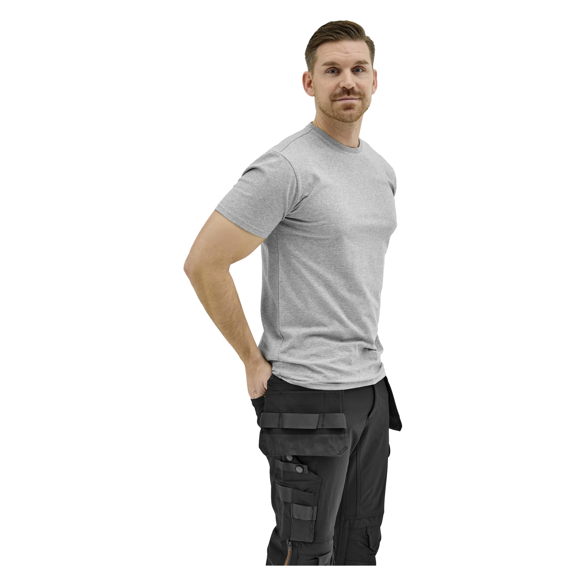 En man med kort hår och mustasch reser sig, klädd i en ljusgrå t-shirt och svarta cargobyxor, ena handen i fickan, och tittar åt sidan.