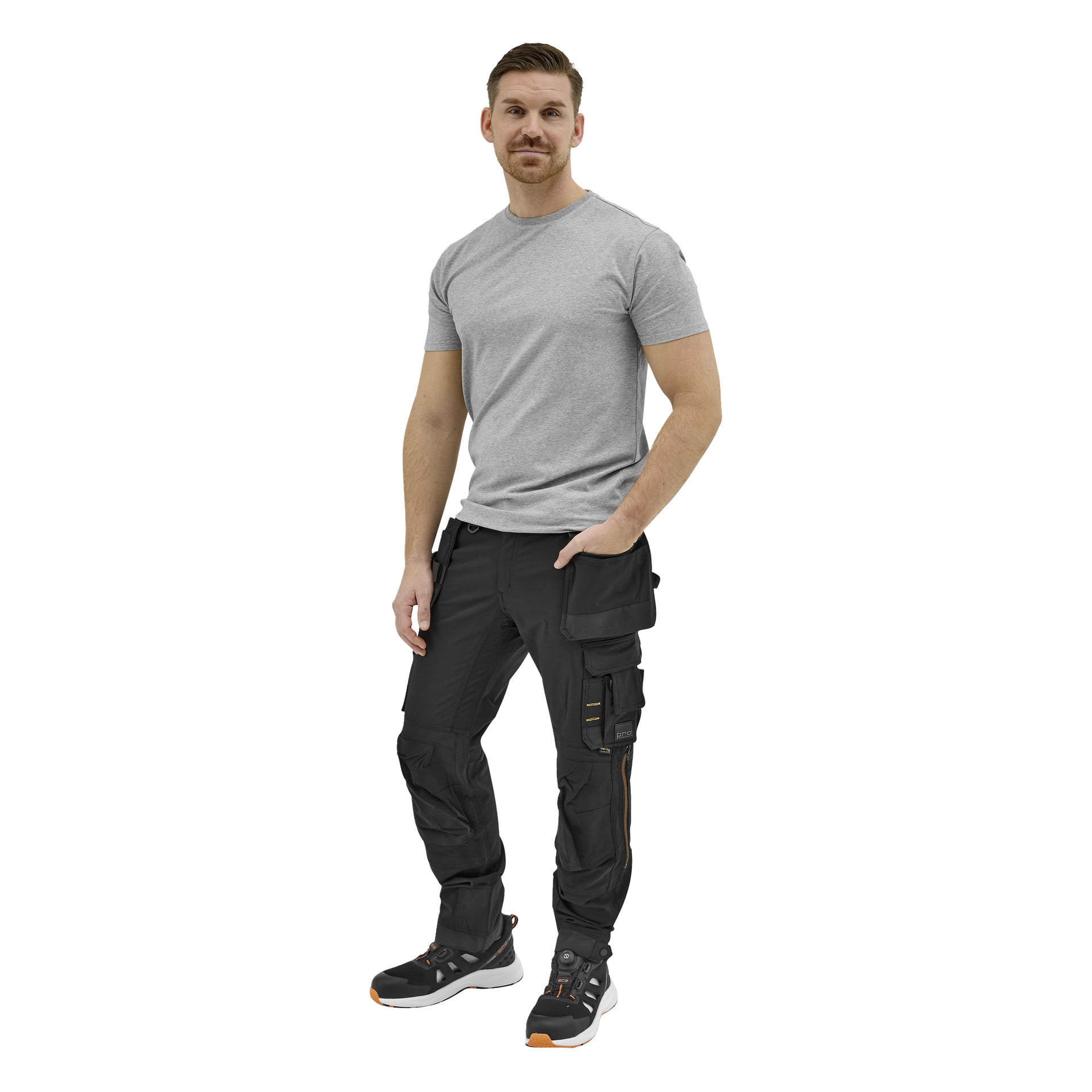 En man står framåt i en ljusgrå kortärmad t-shirt, svarta arbetsbyxor med fickor och svarta sportskor med orange detaljer; en hand i fickan.