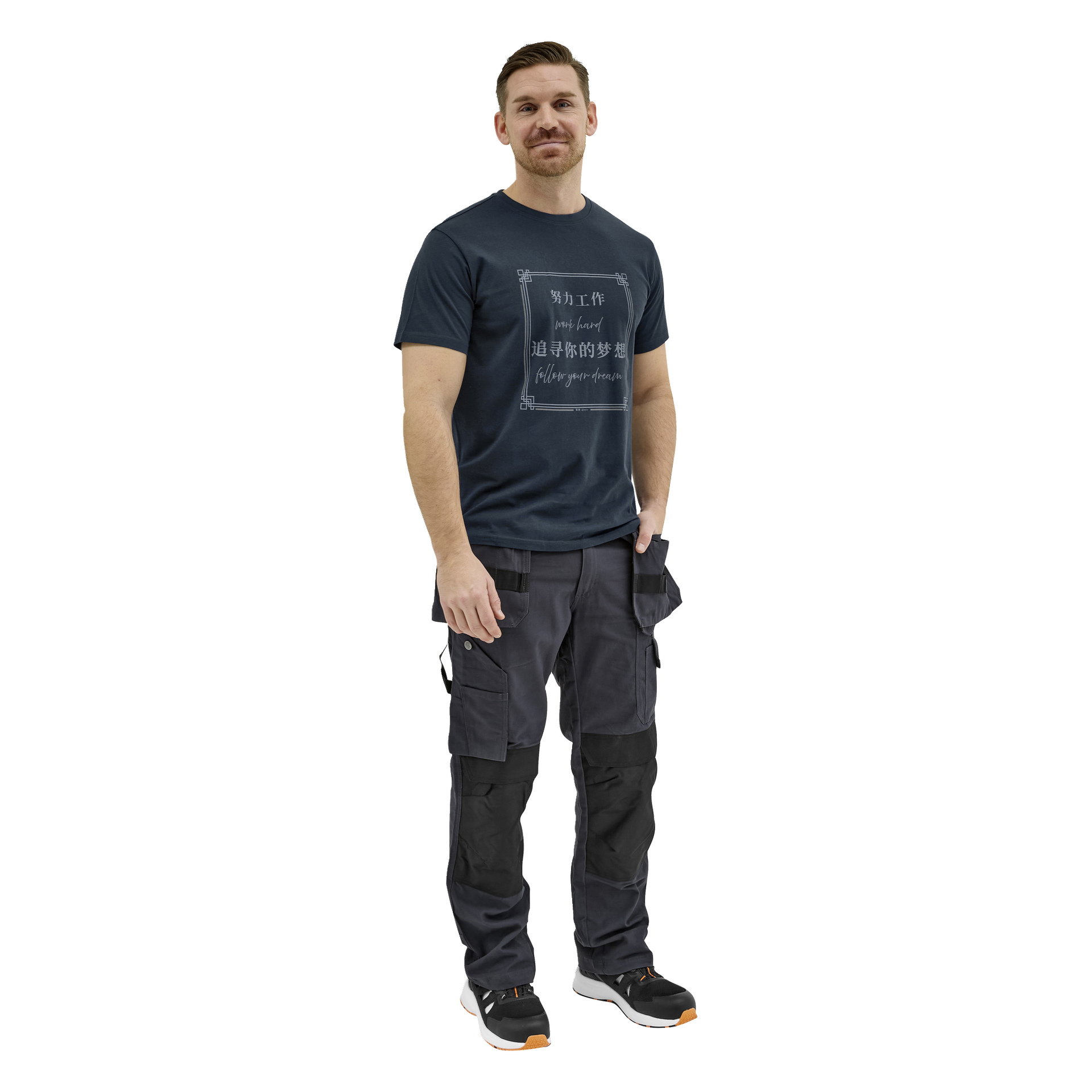 En mann med kort hår og velstelt skjegg står smilende, kledd i en mørk t-skjorte med grafikk, cargobukser med sidelommer og svarte joggesko.
