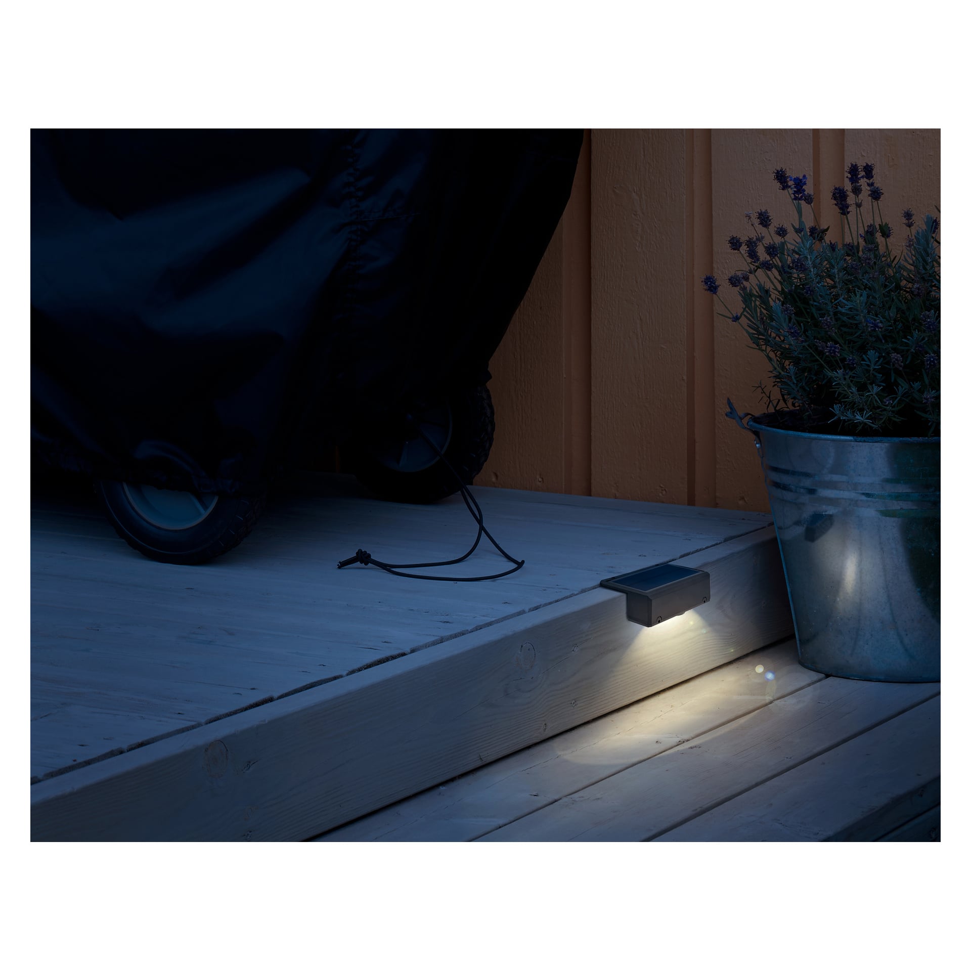 En kompakt rektangulär utomhuslampa på ett trästeg lyser ner. I närheten finns en föremålsdel med hjul och en metallhink med lavendel.