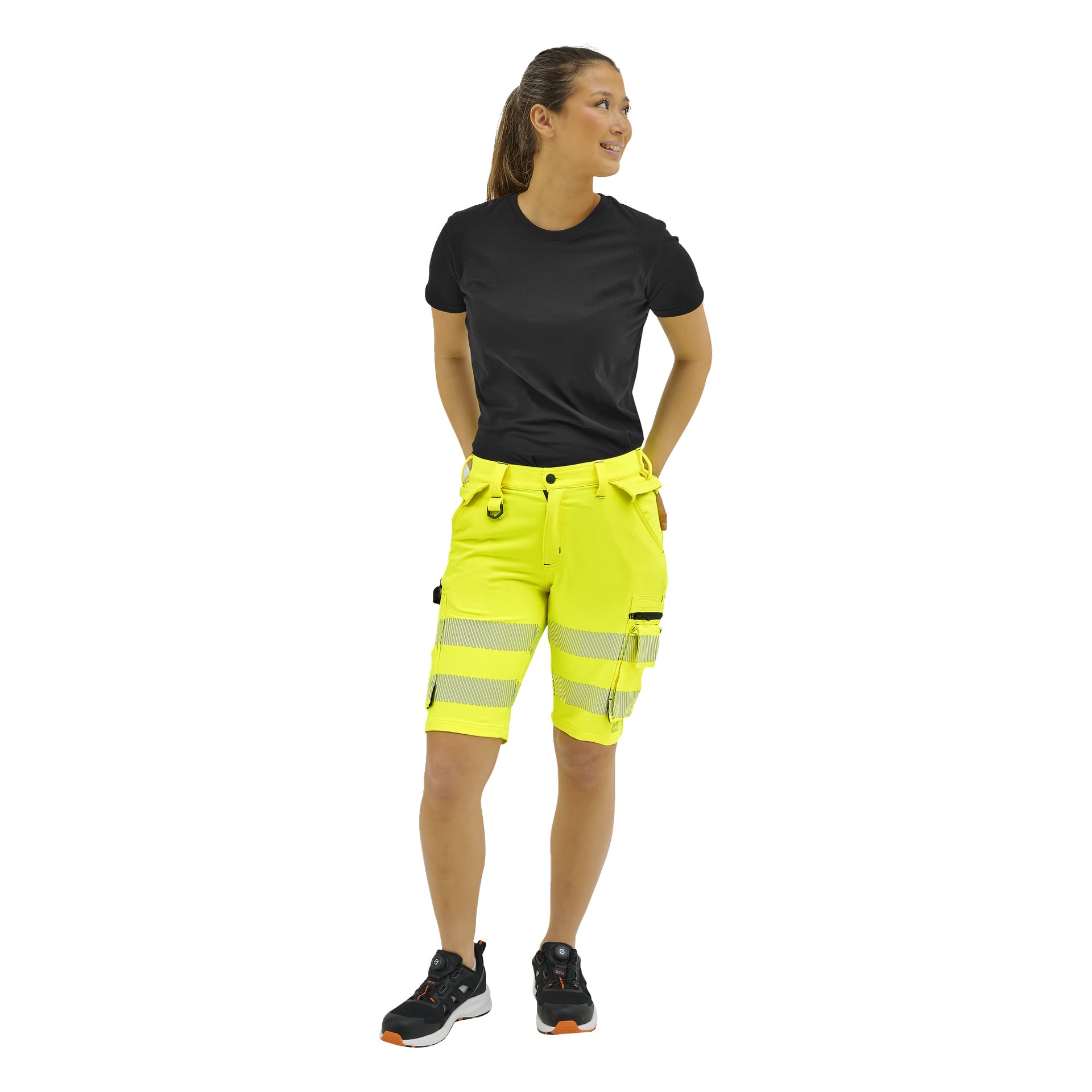 En kvinna som vänder sig åt vänster bär en svart t-shirt, neongula reflekterande arbetsshorts med fickor, svarta arbetsskor och hästsvans.