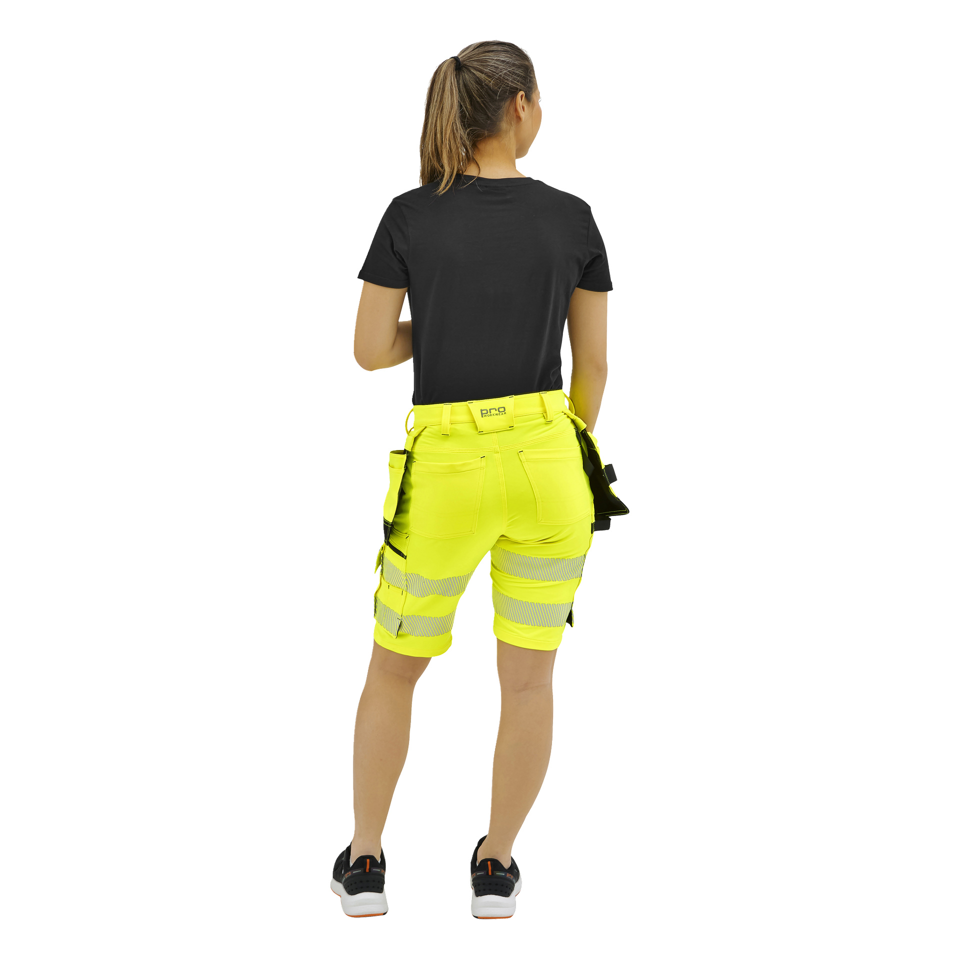 En person med hästsvans står vänd bort, klädd i svart T-shirt, neongula shorts med verktyg och svartvita sneakers.