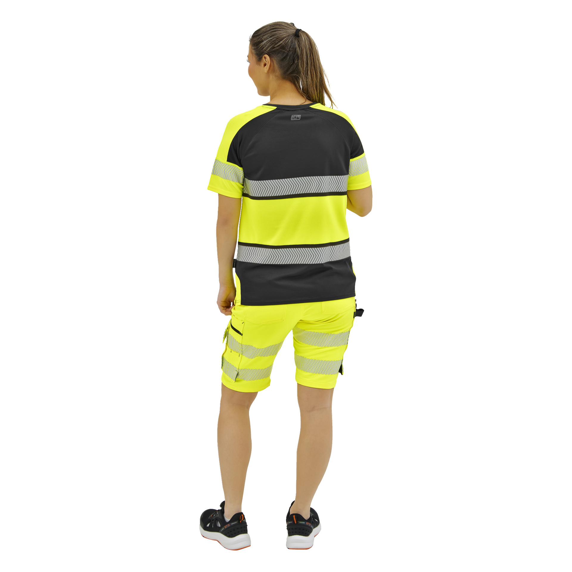 En person står med ryggen vänd, klädd i en neongul och svart atletisk outfit med reflekterande ränder och svarta sneakers.