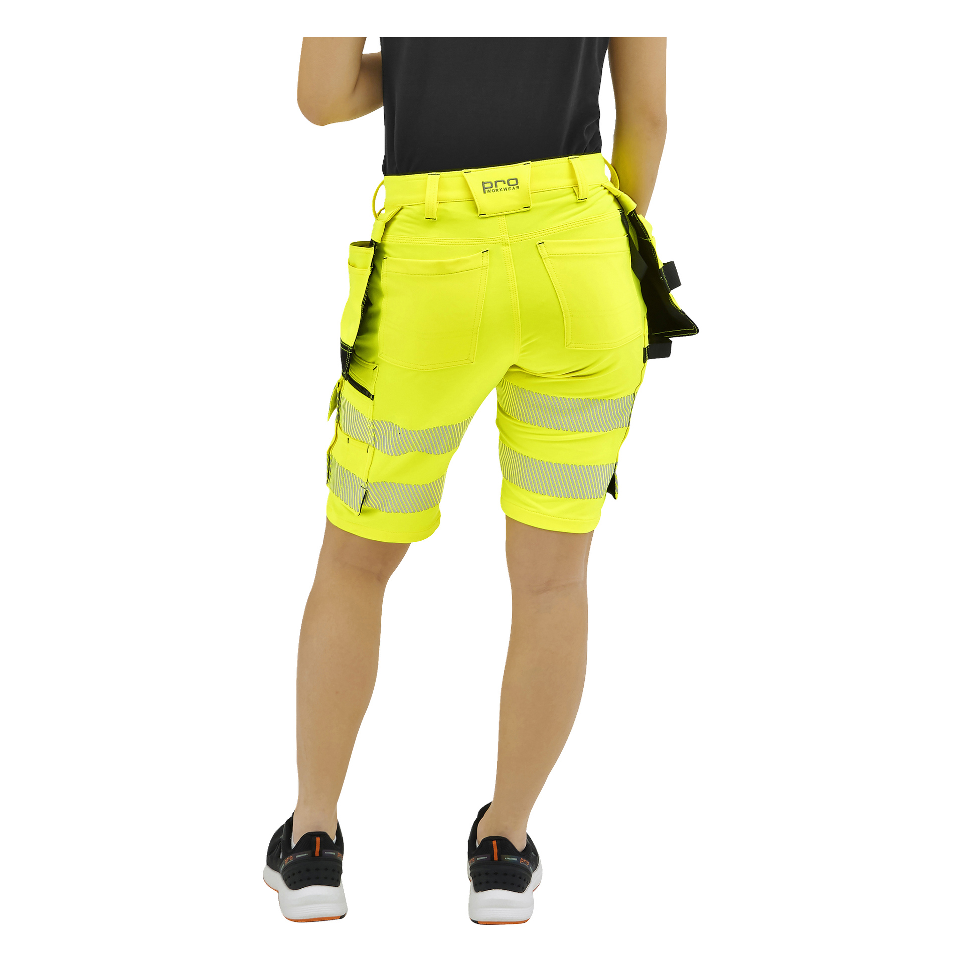 En person som ses bakifrån bär klargula högtidsshorts med reflekterande remsor, svarta fickor, en svart skjorta och svarta sneakers.