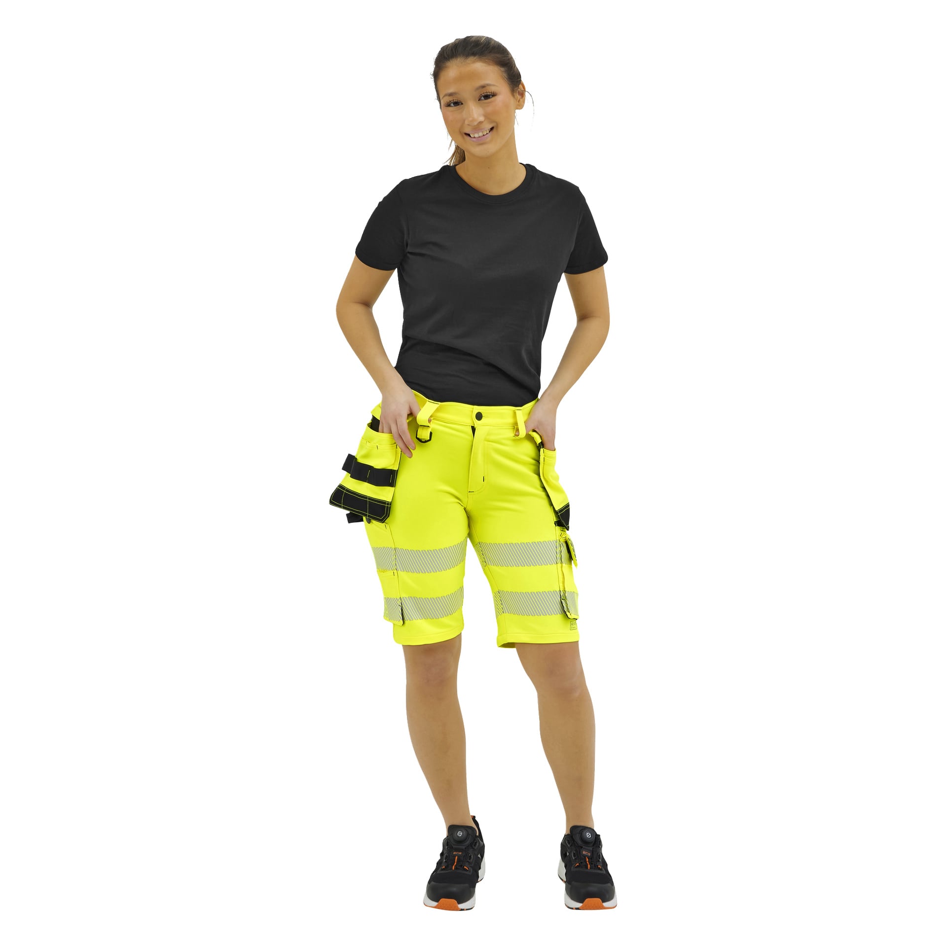 En leende person bär en svart t-shirt, neongula arbetsshorts med reflekterande ränder, svarta arbetsskor och en verktygsväska.