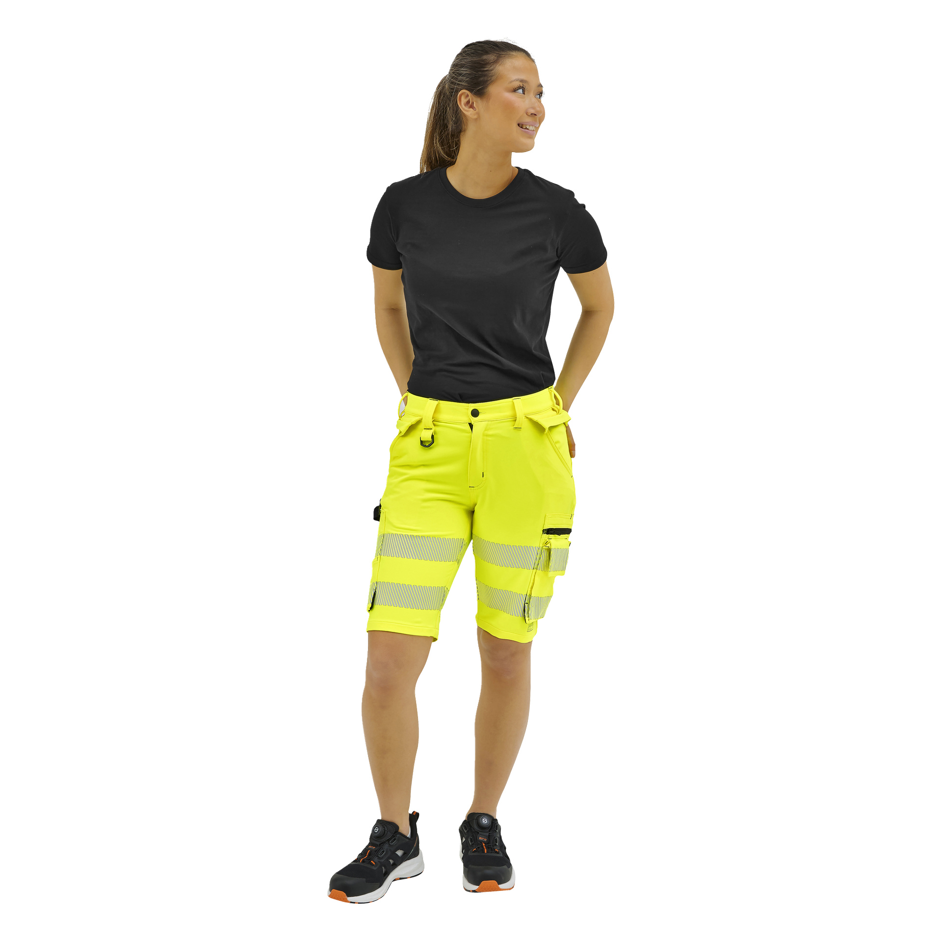 En kvinna, med händerna bakom ryggen, bär en svart t-shirt, gula högtidsshorts med reflekterande ränder, svarta strumpor och arbetsskor.