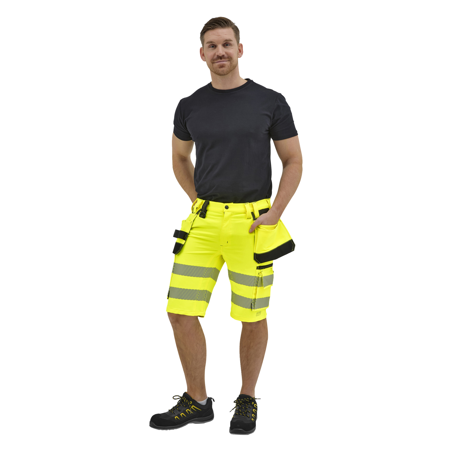 En man står vänd framåt i en svart t-shirt och neongula arbetsshorts med reflekterande ränder, svarta skor med gula detaljer, vänster hand i fickan.