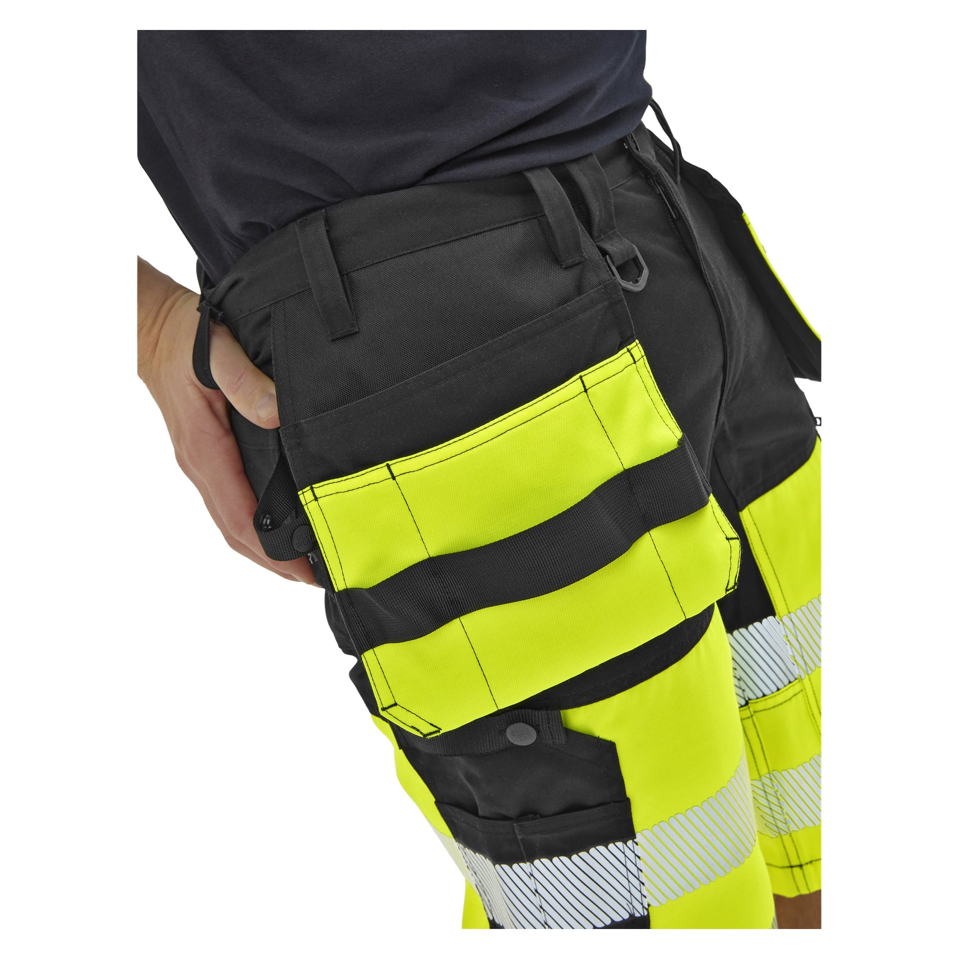En person i svarta och gula hög-vis-arbetsbyxor med reflekterande ränder och flera fickor, visade från midja till mitten av låret, hand i ficka.