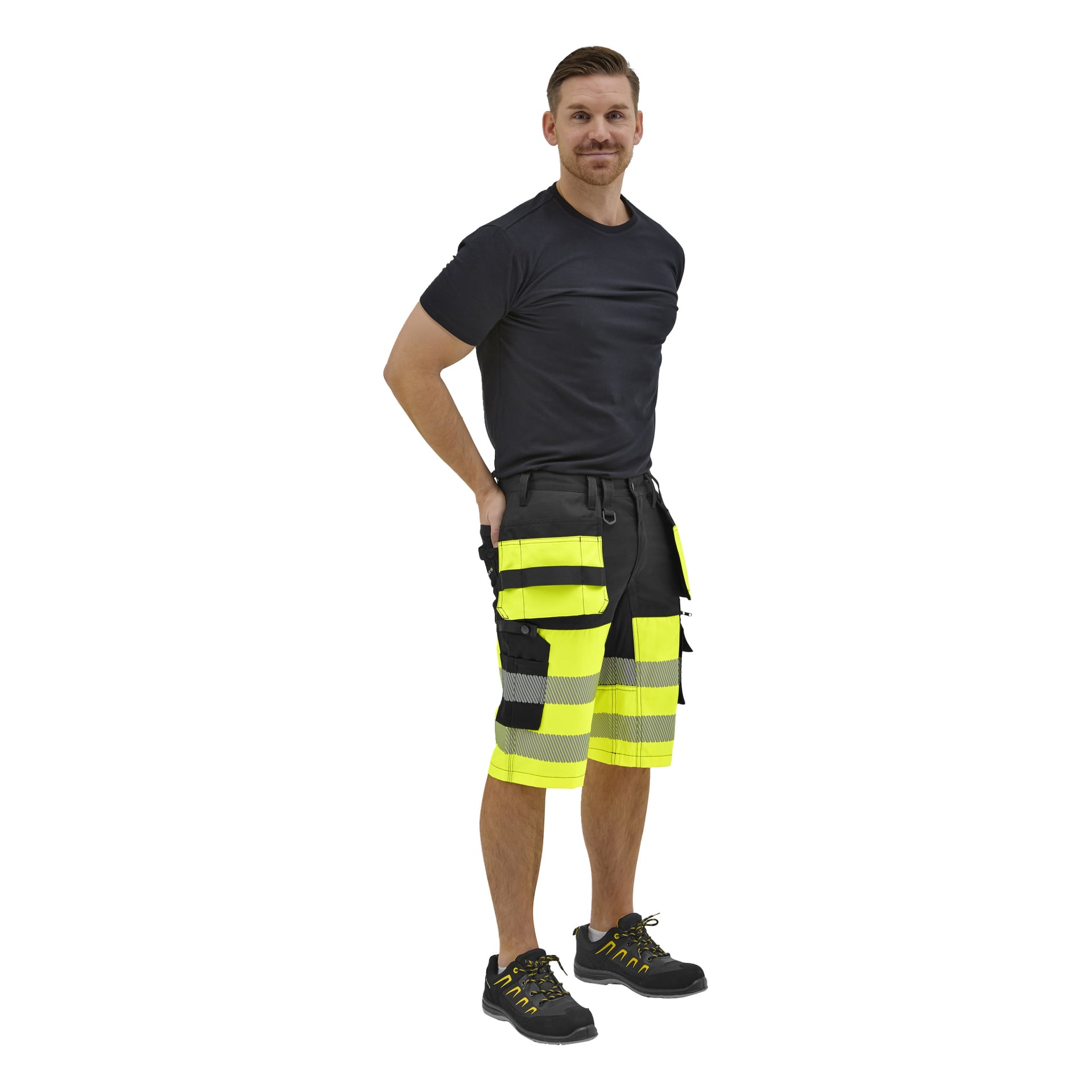 En man står lätt vänd åt sidan i en svart t-shirt, svarta och gula cargoshorts med hög synlighet och svarta skor med gula detaljer.