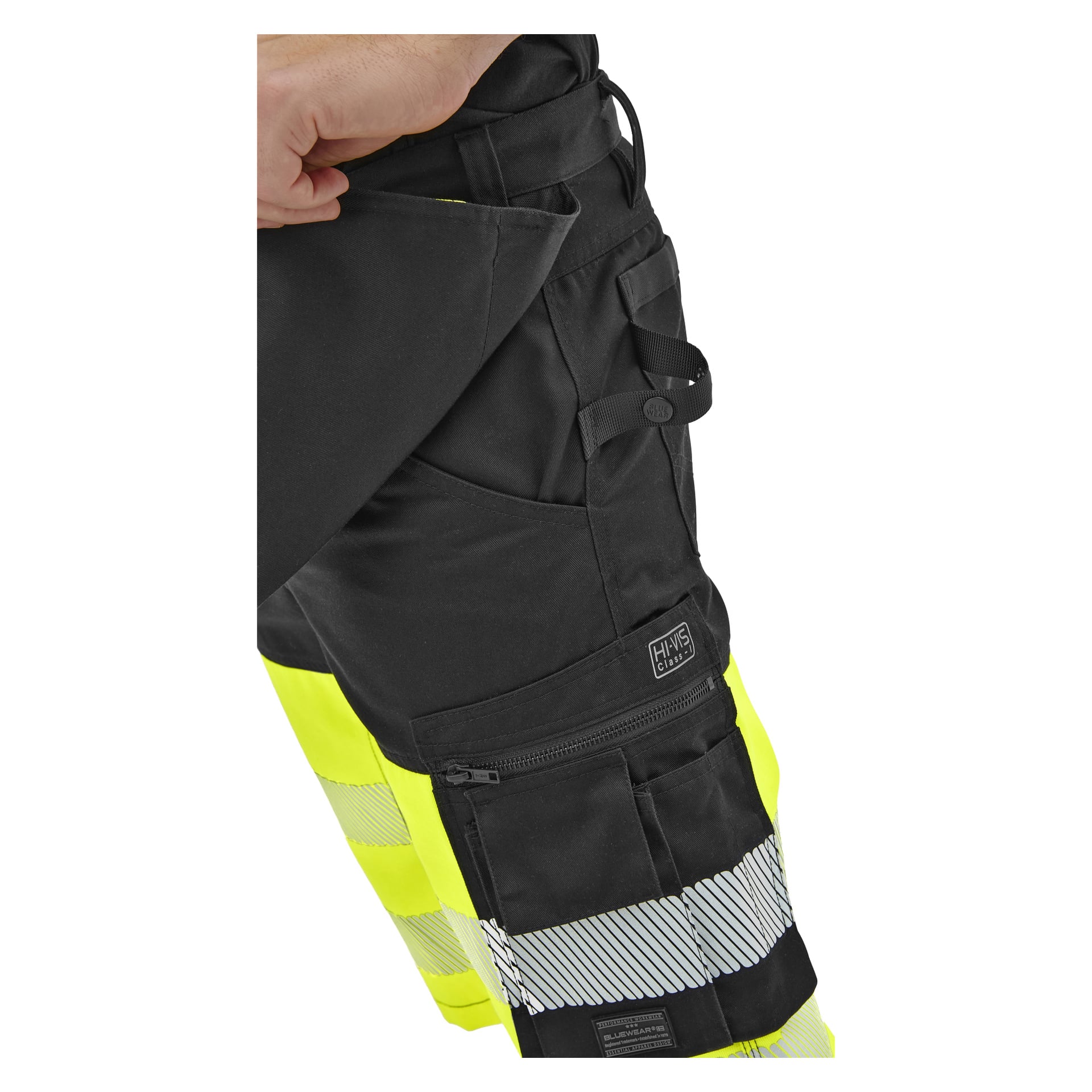 Närbild på svarta och gula hi-vis arbetsbyxor med reflekterande remsor när en hand lyfter en flik nära fickan och avslöjar sömdetaljer.