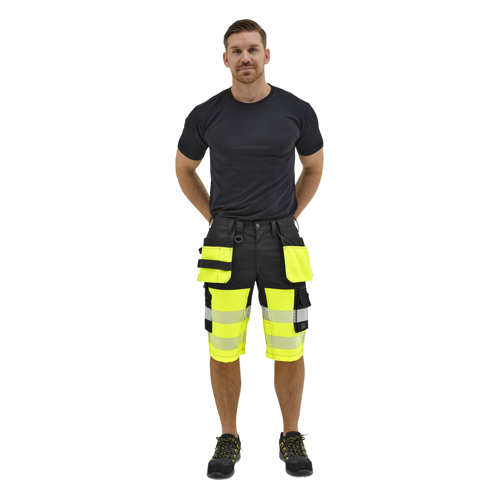 En man står framåt, klädd i svart T-shirt, neongula och svarta högprofilerade arbetsshorts med reflekterande ränder och svarta skor med gula snören.