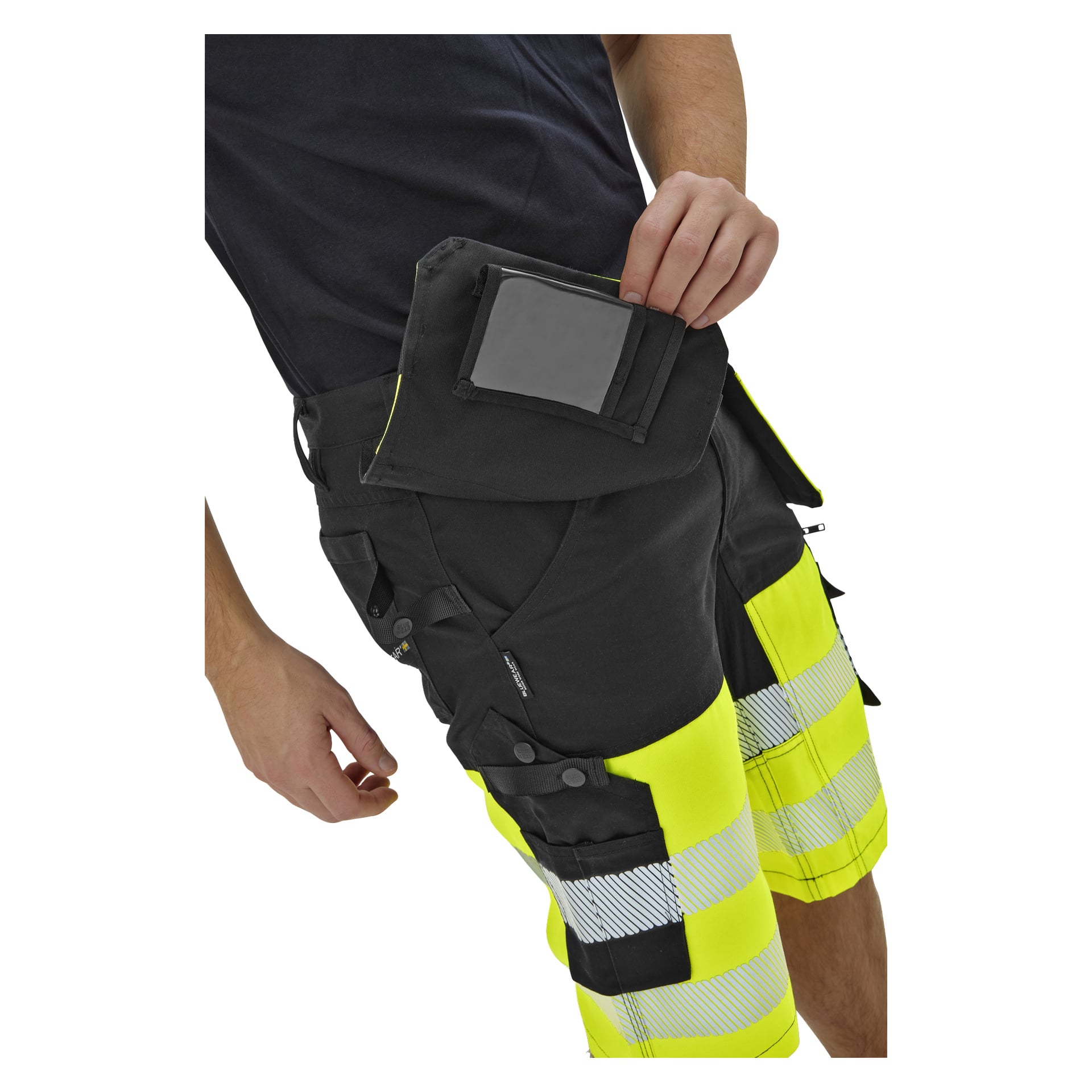 En person i svarta och gula högtidsshorts med reflekterande ränder stoppar en svart plånbok eller påse i en ficka; bål och ben synliga.