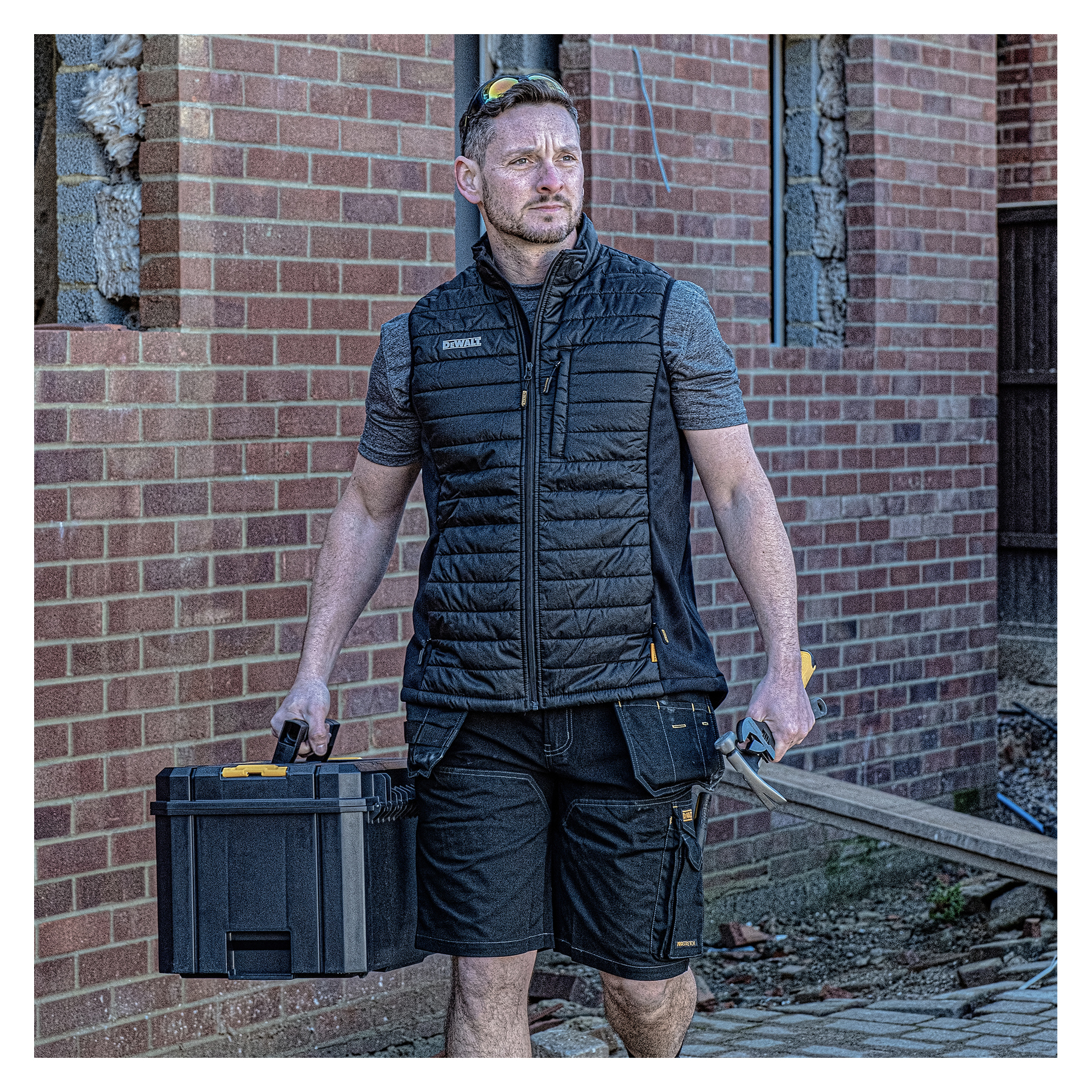 Ein Mann in schwarzer Weste und Shorts geht an einem im Bau befindlichen Backsteingebäude vorbei, hält einen schwarzen Werkzeugkasten und eine Handsäge in der Hand.