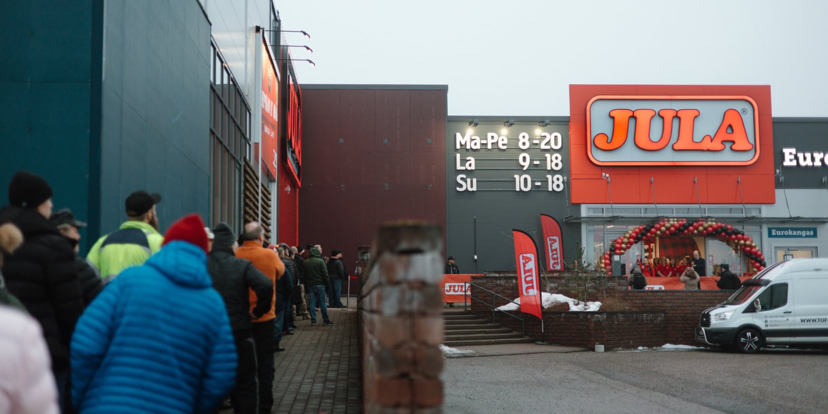 Talvitakkeissa ihmiset odottavat Jula-kaupan ulkopuolella, jonka sisäänkäynnillä on punaisia kylttejä, punaisia lippuja ja ilmapalloja. Valkoinen pakettiauto on pysäköity lähelle.