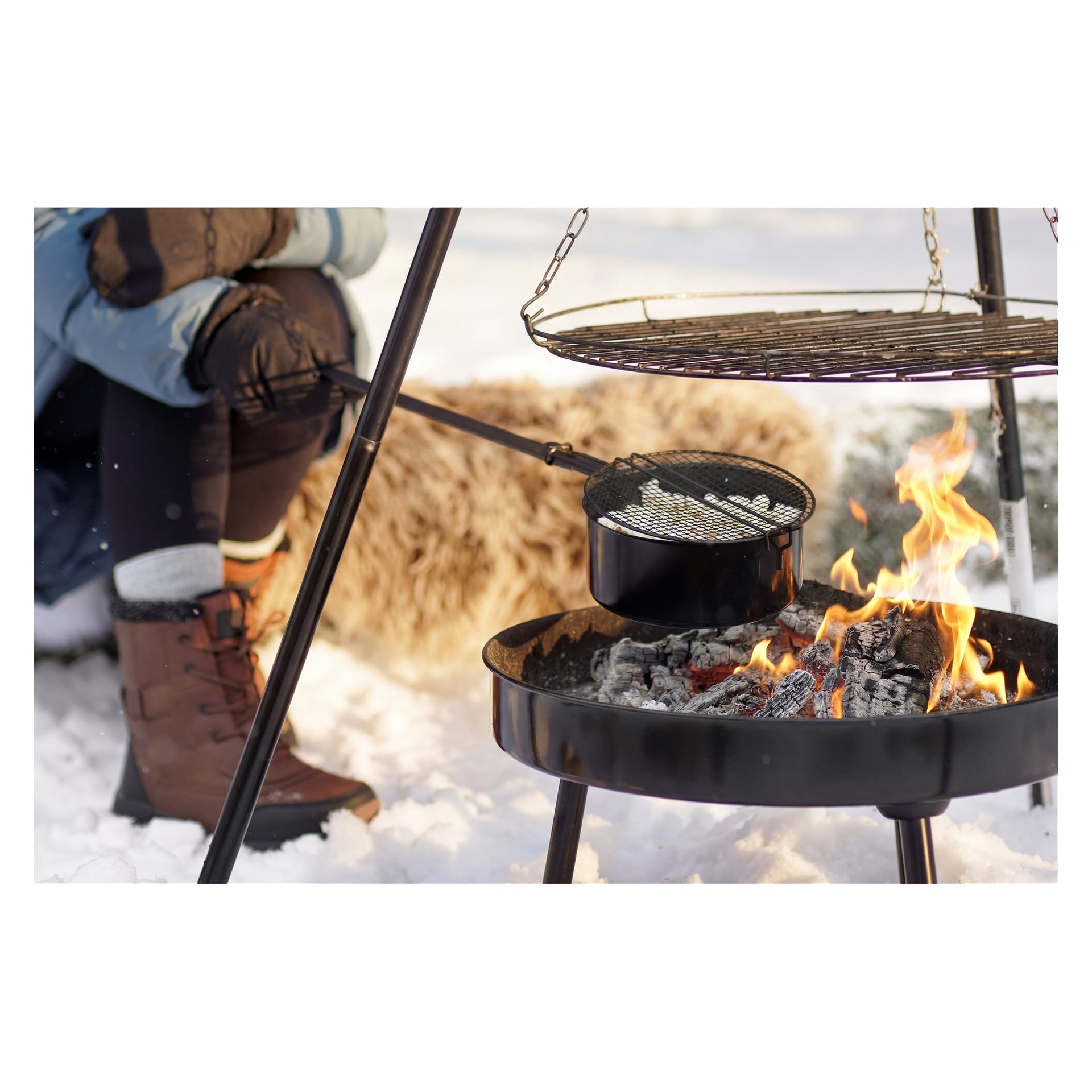 Inlindad i vinterstövlar och en puffjacka lagar någon mat över en snötäckt eldstad med en panna, grill och en lurvig filt i närheten.
