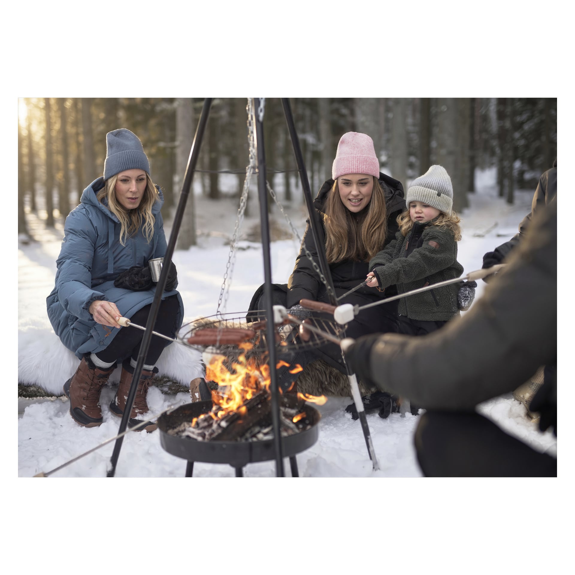Tre vuxna och ett barn i vinterkläder sitter i snön vid en eldstad och rostar korvar, omgivna av snötäckta skogsträd.
