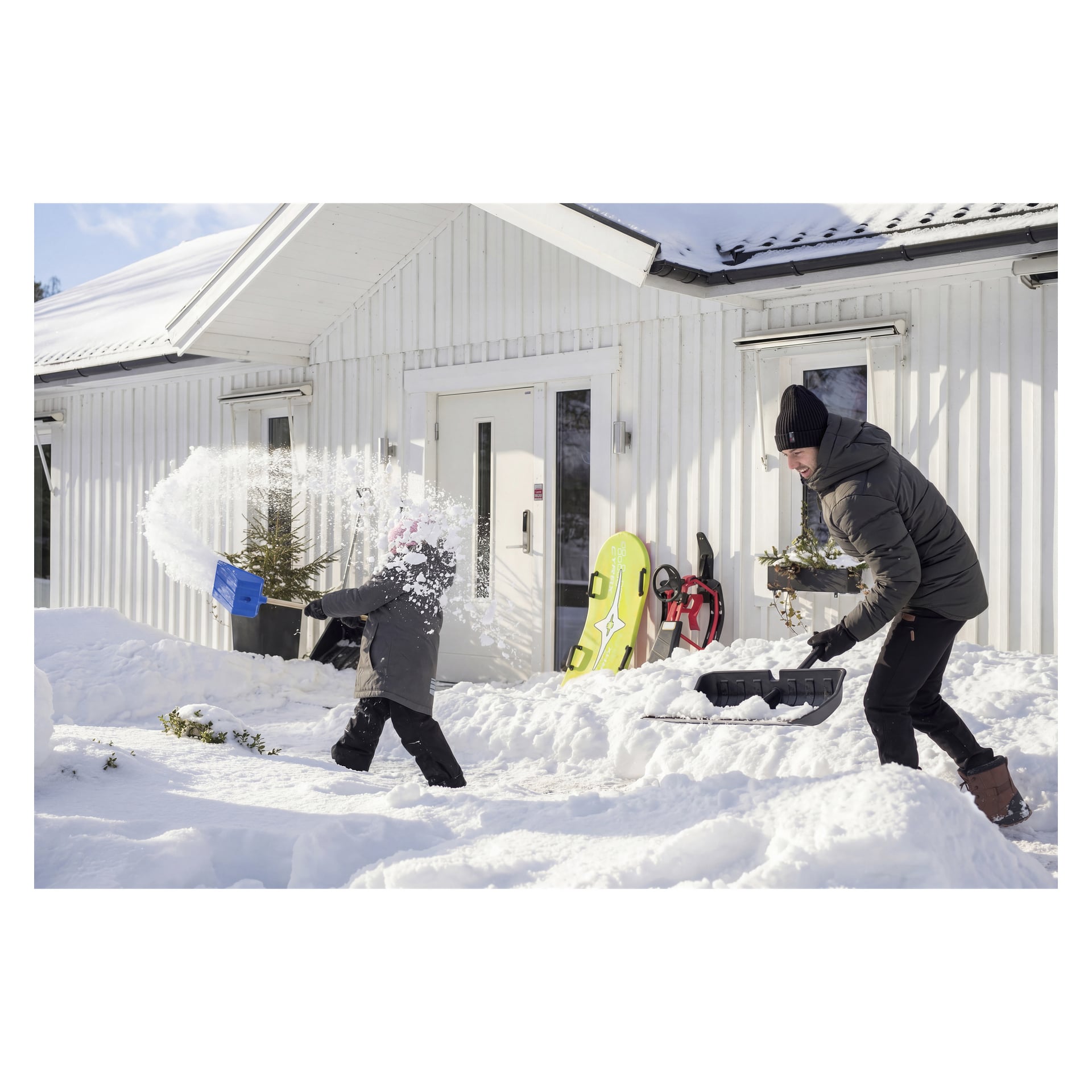 En vuxen och ett barn skyfflar snö vid ett hus; barnet använder en blå spade, den vuxna håller en svart. Slädar vilar i närheten.