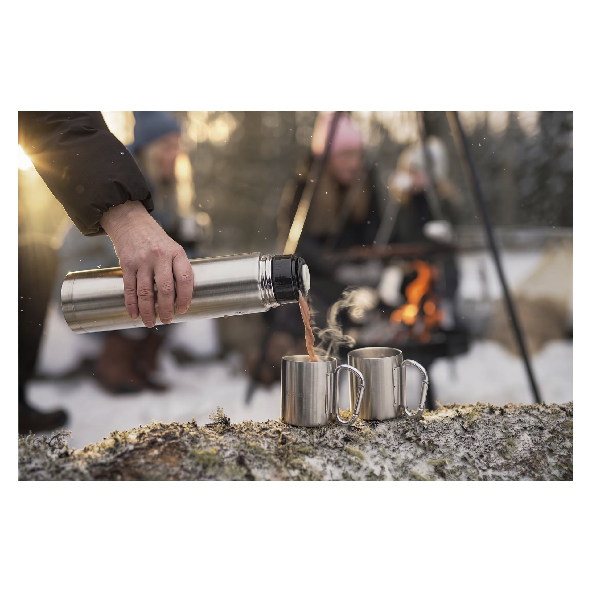 Någon häller upp en ångande dryck ur en termos i två metallmuggar på en stock, medan andra samlas vid en lägereld i snötäckta skogar.