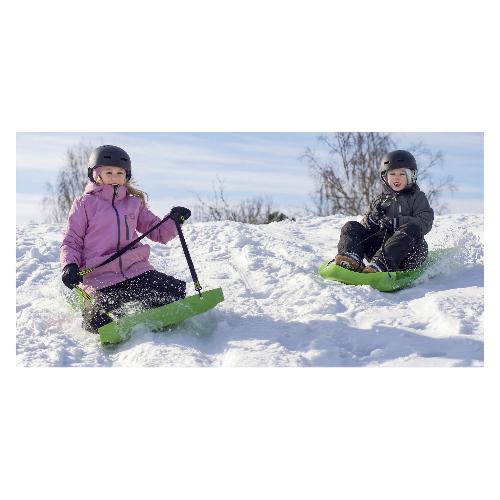 Två barn i hjälmar och vinterkläder åker släde nerför en snötäckt kulle på gröna slädar, ett i rosa, ett i mörka kläder. Nakna träd bakom.