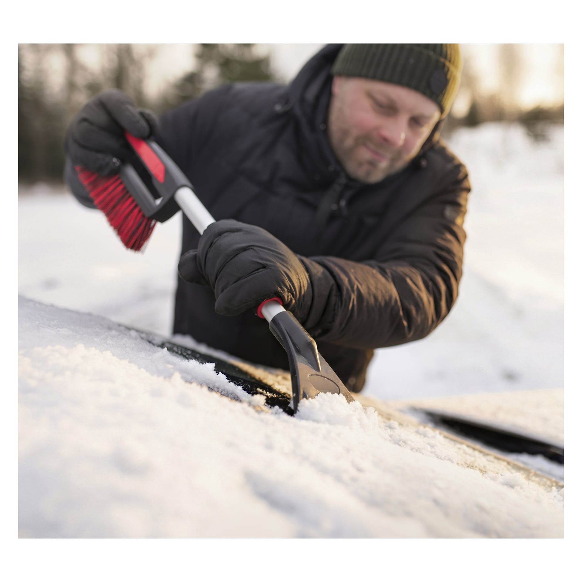 Någon i vinterjacka, handskar och mössa använder en skrapa för att rensa snö från en bilruta utomhus i snön.