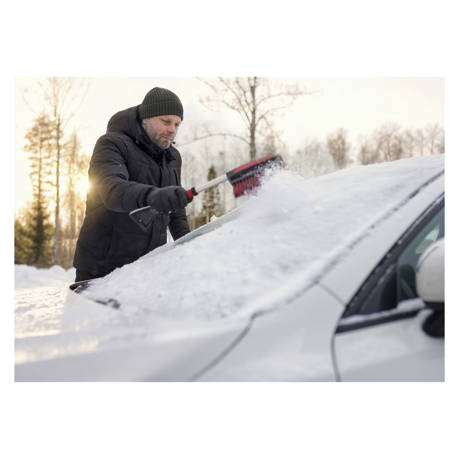 En man i svart jacka och grön hatt borstar snö från en vit bils vindruta en snöig dag, med träd och solljus bakom sig.