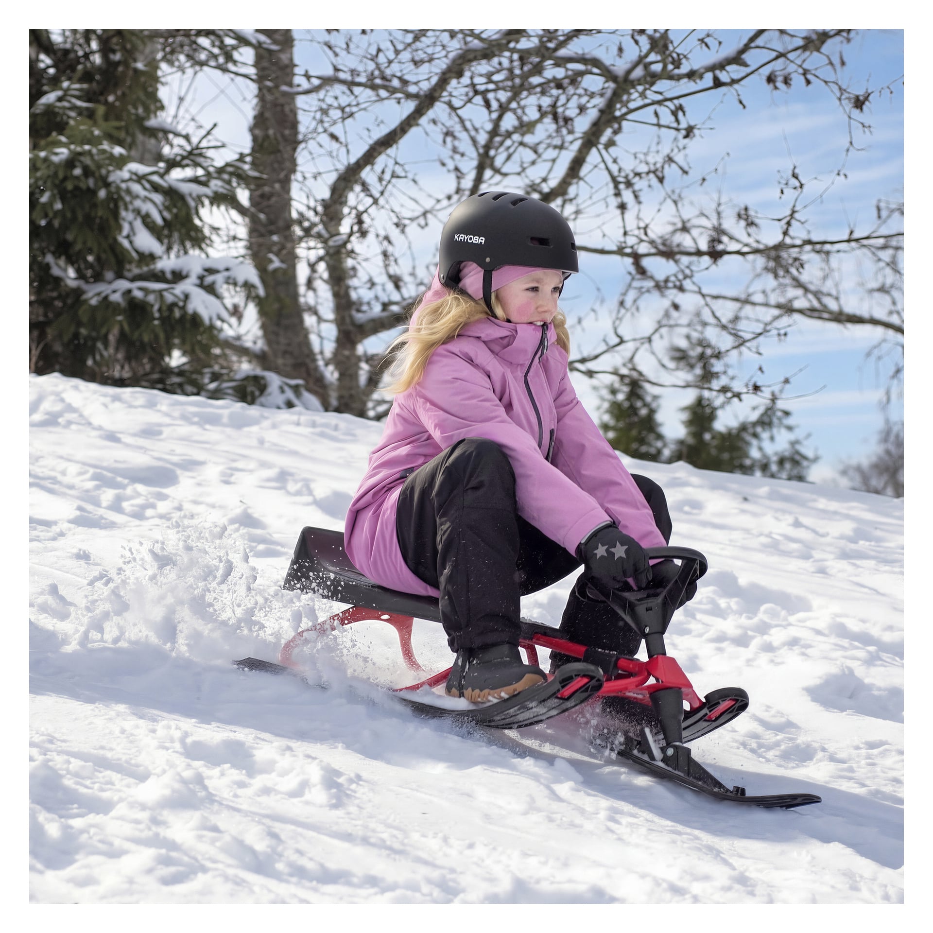 Ett barn i hjälm, rosa jacka och svarta byxor åker släde nerför en snötäckt kulle på en röd och svart släde med träd bakom.