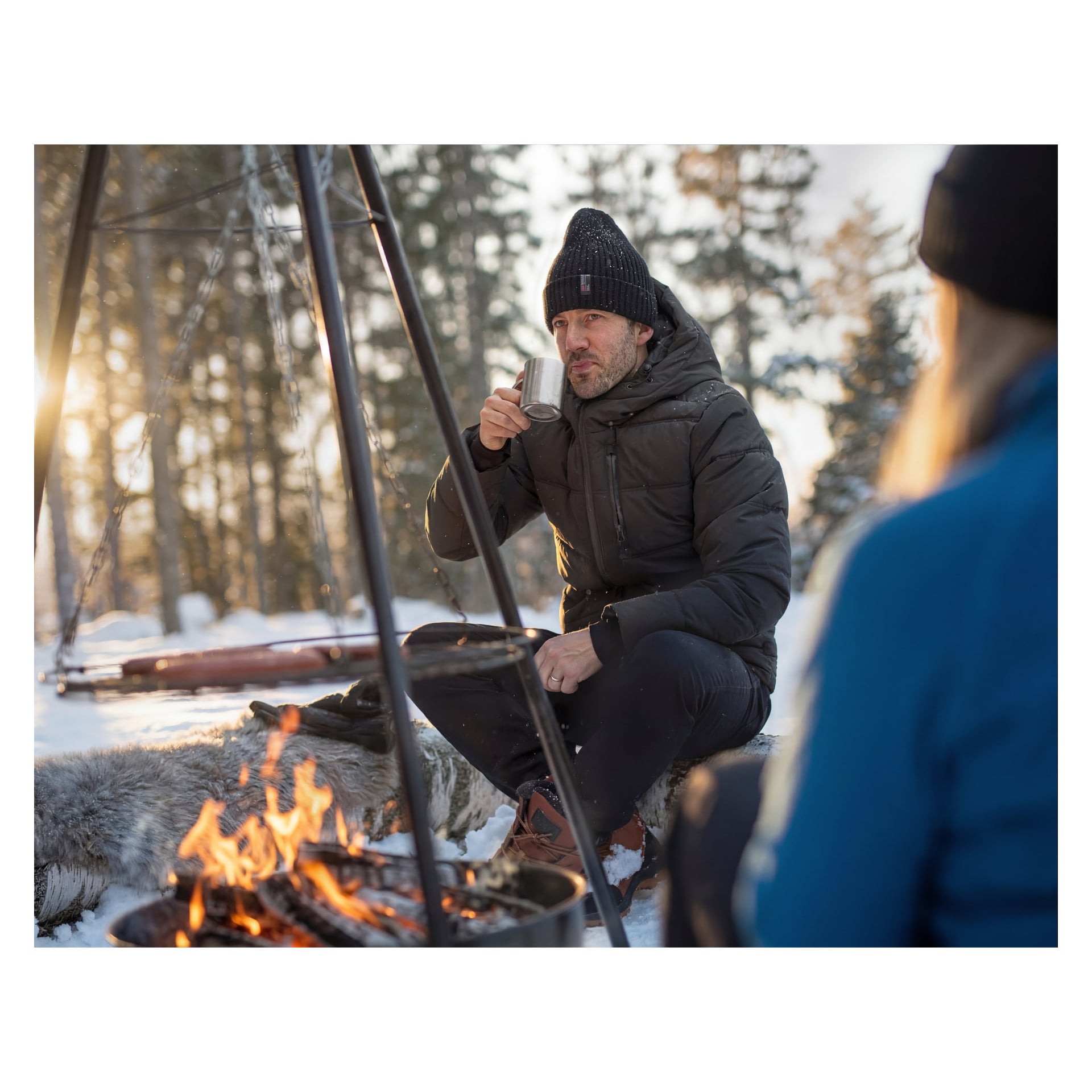 En man i svart jacka och mössa sitter vid en lägereld på snön och håller i en metallmugg. En annan person i blått är i förgrunden.