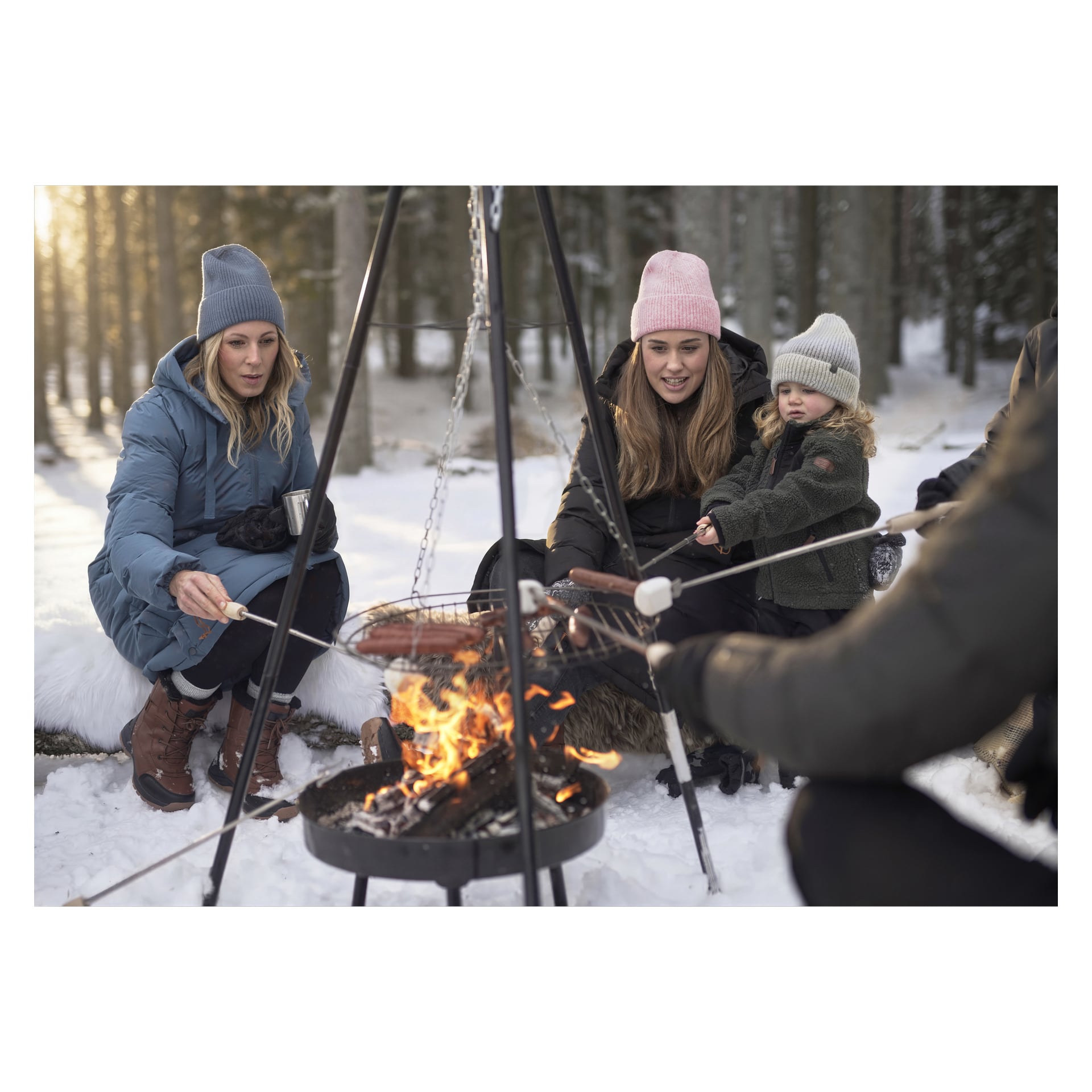 Drei Erwachsene und ein Kind in Winterkleidung rösten Würste über einem Feuer in einem verschneiten Wald; Einer hält eine Tasse, während Schnee und Bäume ihn umgeben.