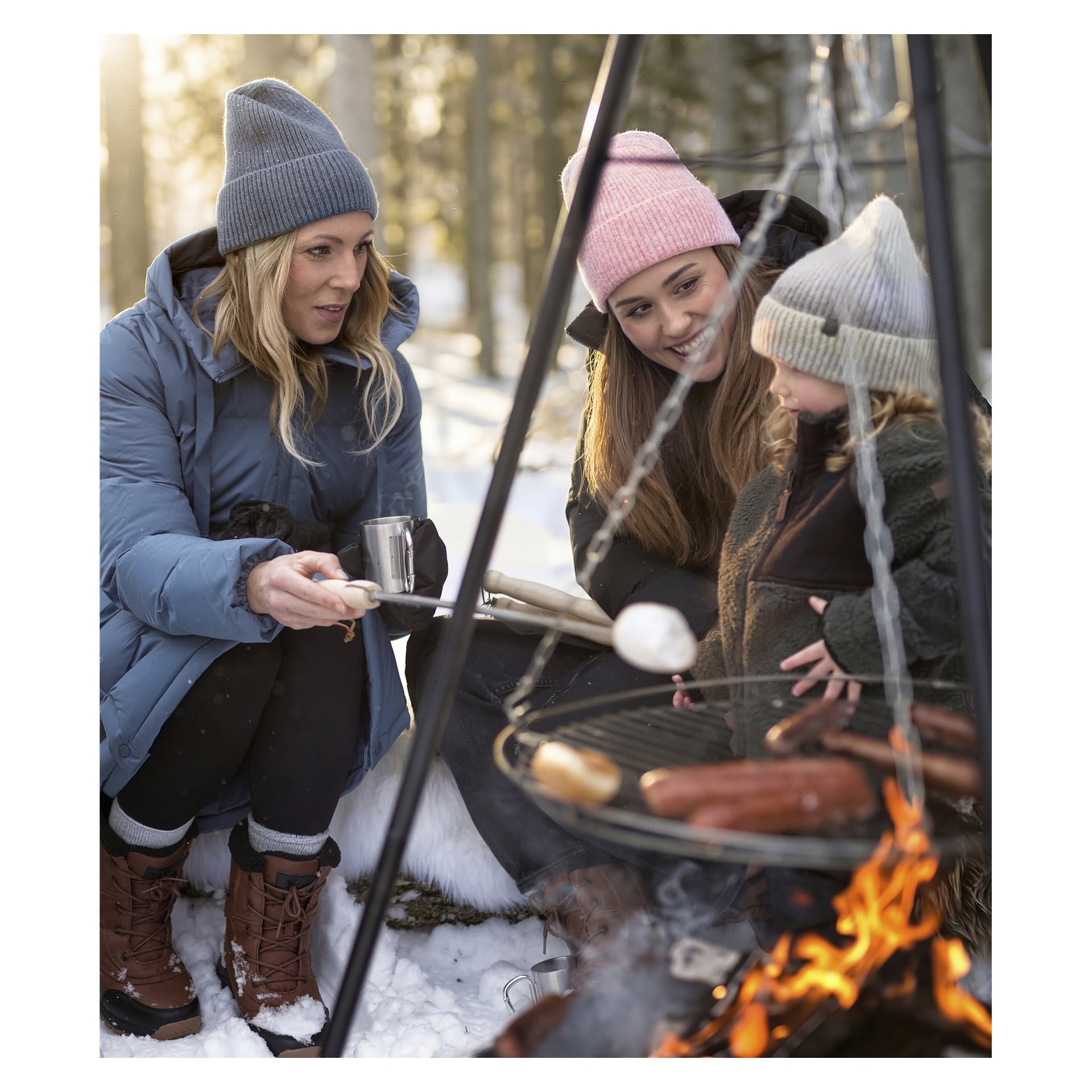 Drei Personen in Winterkleidung versammeln sich im Schnee um ein Lagerfeuer und rösten Marshmallows und Würstchen. Einer hält eine Tasse.