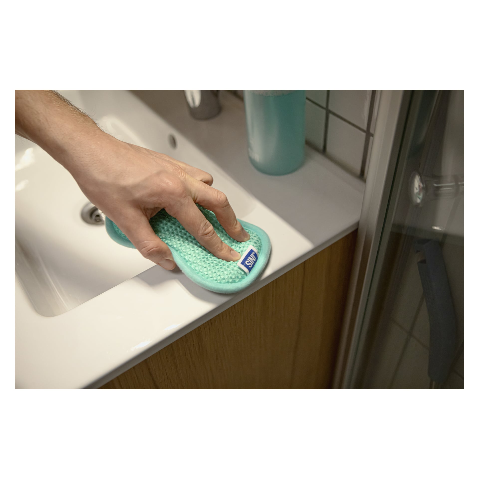 Käsi pyyhkii valkoisen kylpyhuoneen altaan reunaa sinisellä Scrub Daddy -sienellä. Pullo, joka on täytetty sinisellä nesteellä, on tiskialtaan takana.