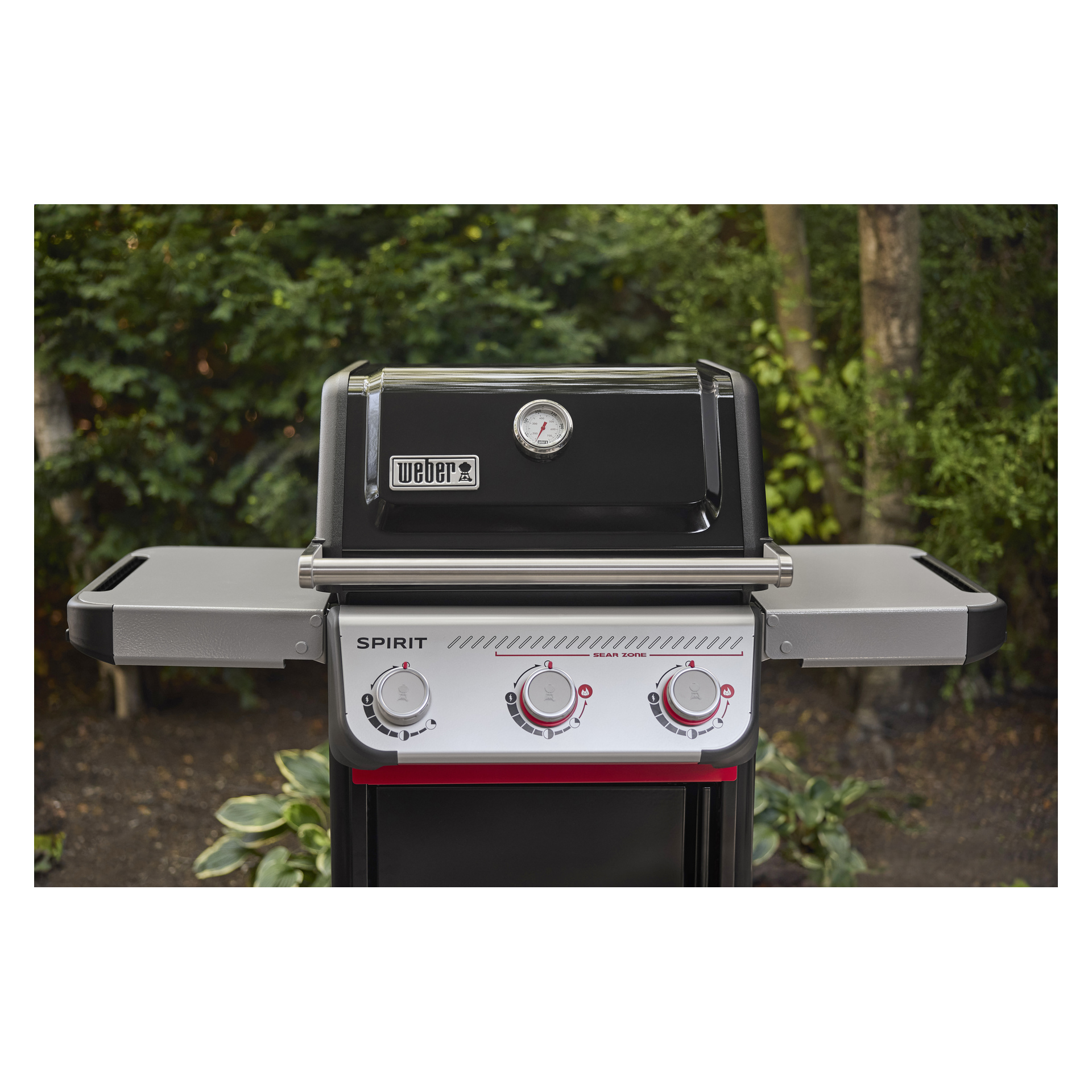 En Weber Spirit gasgrill med stängt svart lock, temperaturmätare, tre mätare och metallhyllor i sidorna, utomhus bland grönska.