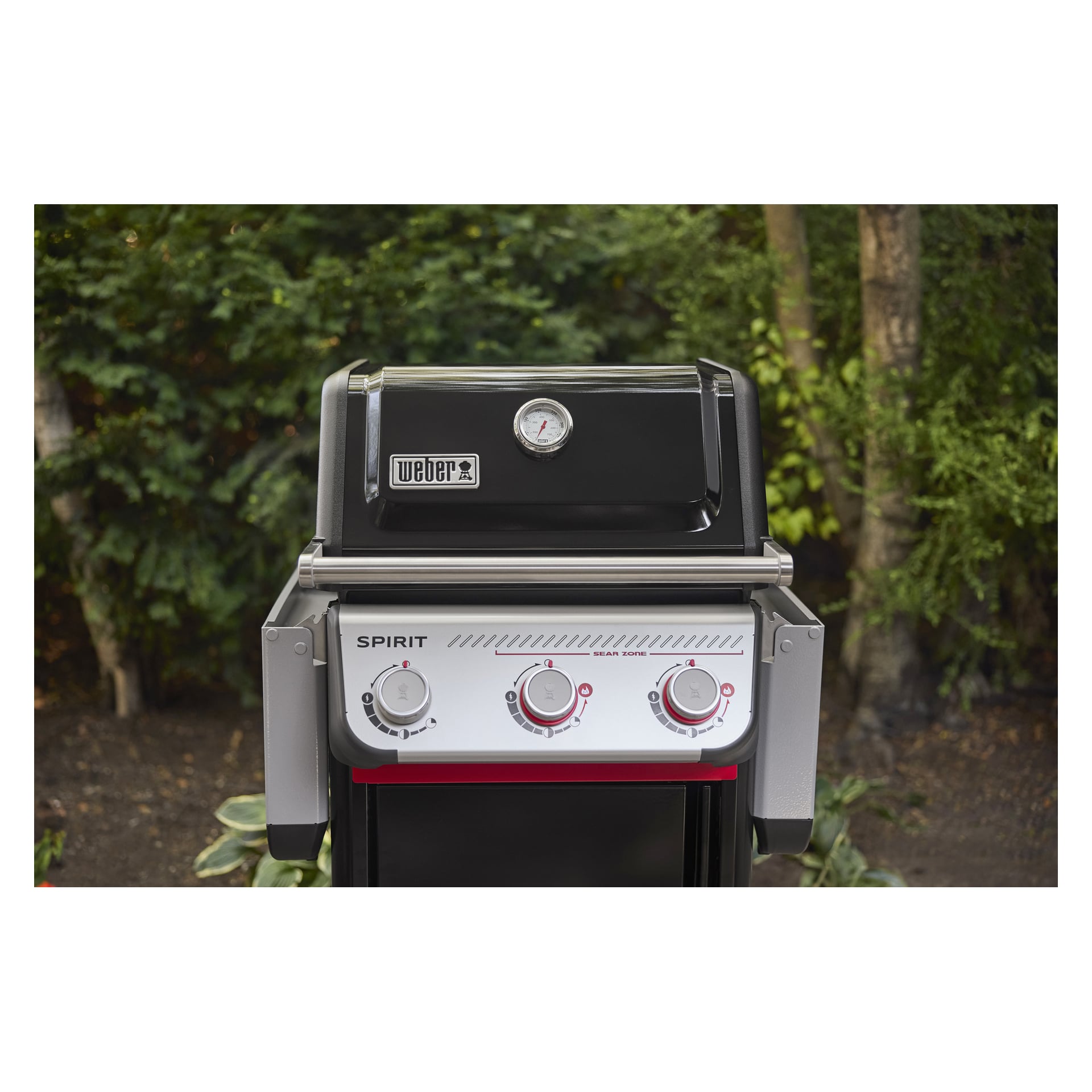 En Weber Spirit gasgrill med locket stängt och tre vred står utanför, omgiven av frodiga gröna växter och träd.
