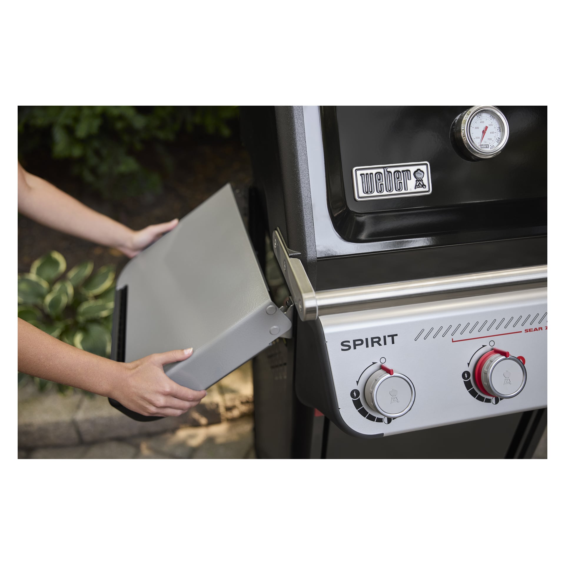Någon sänker sidohyllan på en svart Weber Spirit gasgrill med kontrollrattar och en inbyggd termometer.