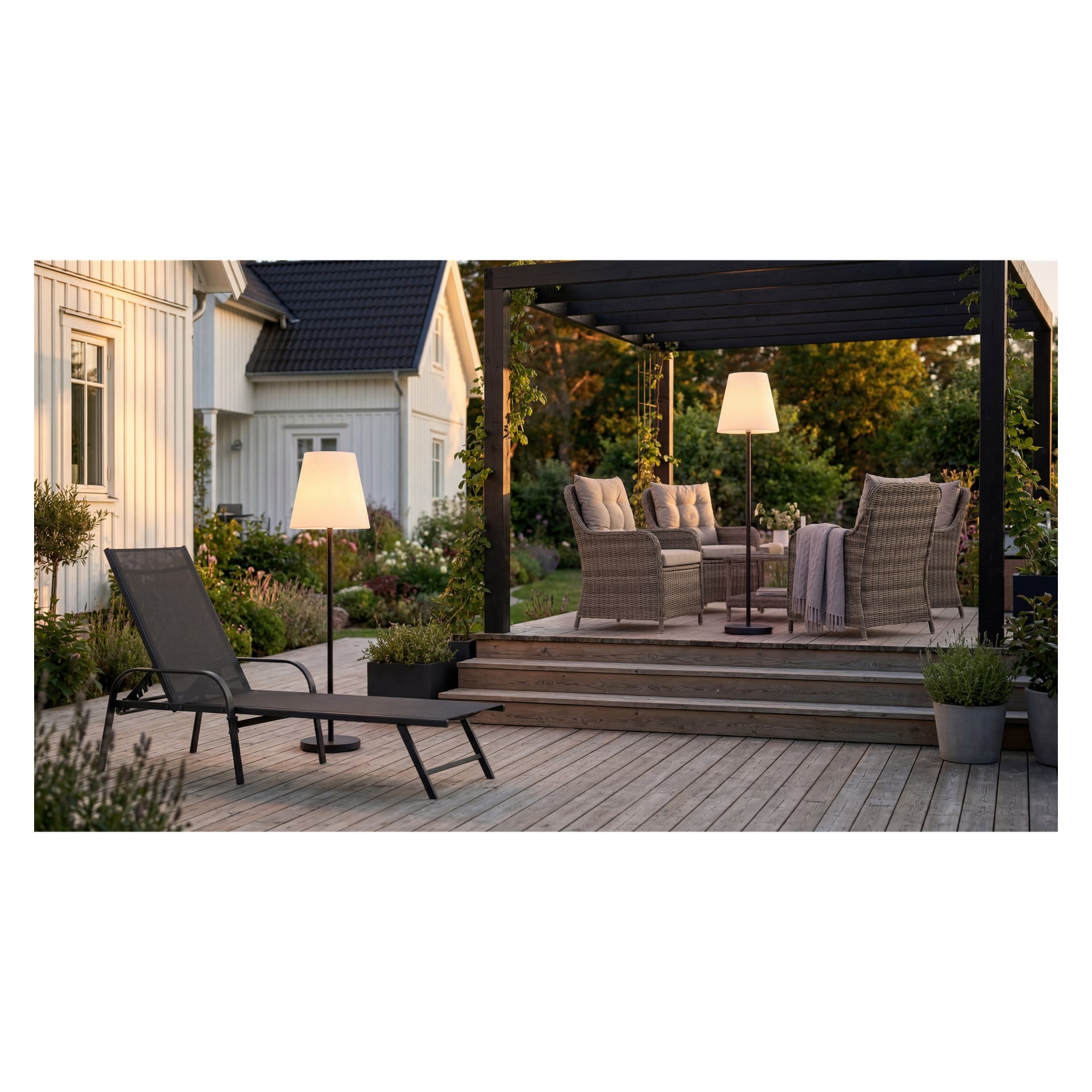Holzterrasse mit Weidenstühlen, ein Tisch unter der Pergola, Stehlampen, ein Liegestuhl, Grünflächen und ein weißes Haus im Hintergrund.