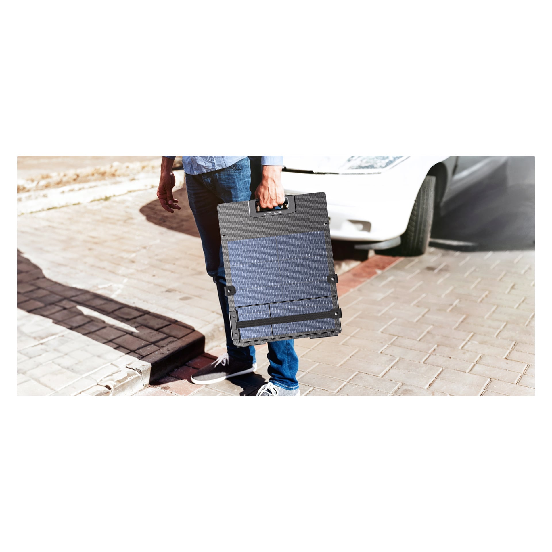 Någon på en asfalterad trottoar håller en rektangulär portabel solpanel i handtaget, iklädd jeans och sneakers; En vit bil är bakom.