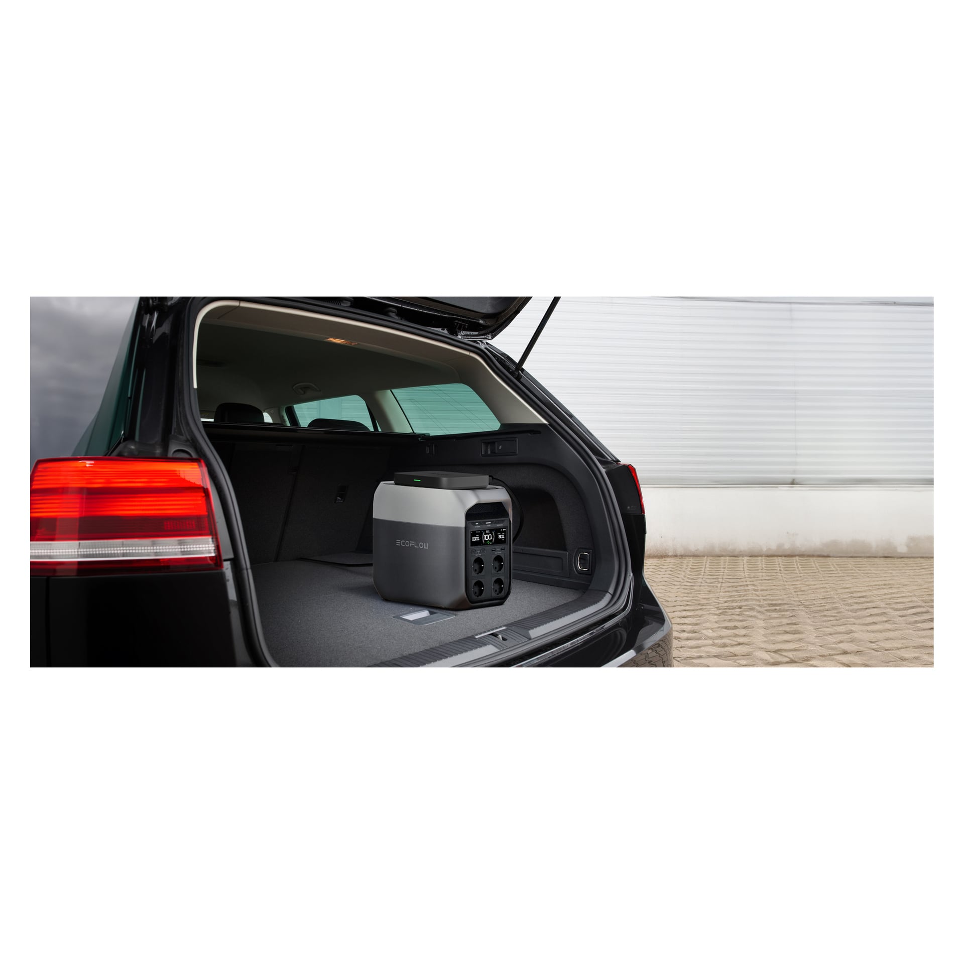 Ett portabelt kraftverk står i det öppna bagageutrymmet på en svart SUV parkerad på asfalt, med en vit metallvägg bakom fordonet.