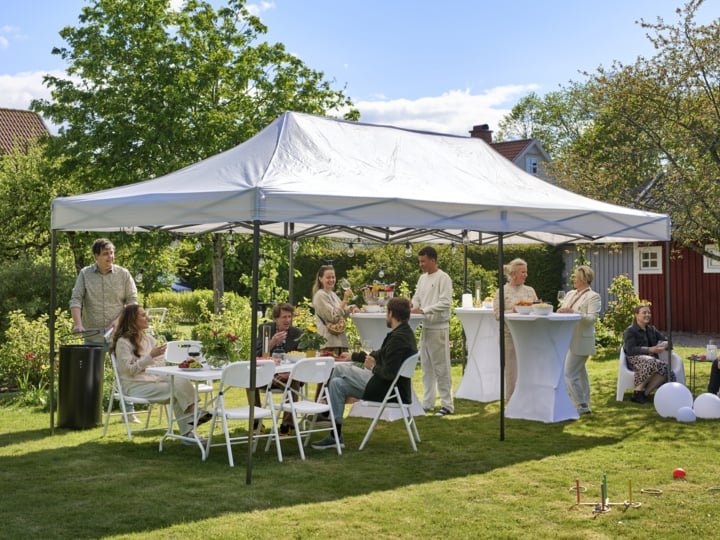 Menschen unterhalten sich in einem grasbewachsenen Garten unter einem großen weißen Blätterdach; Einige sitzen an Tischen, andere stehen in der Nähe von Cocktailtischen zwischen Bäumen und Häusern.