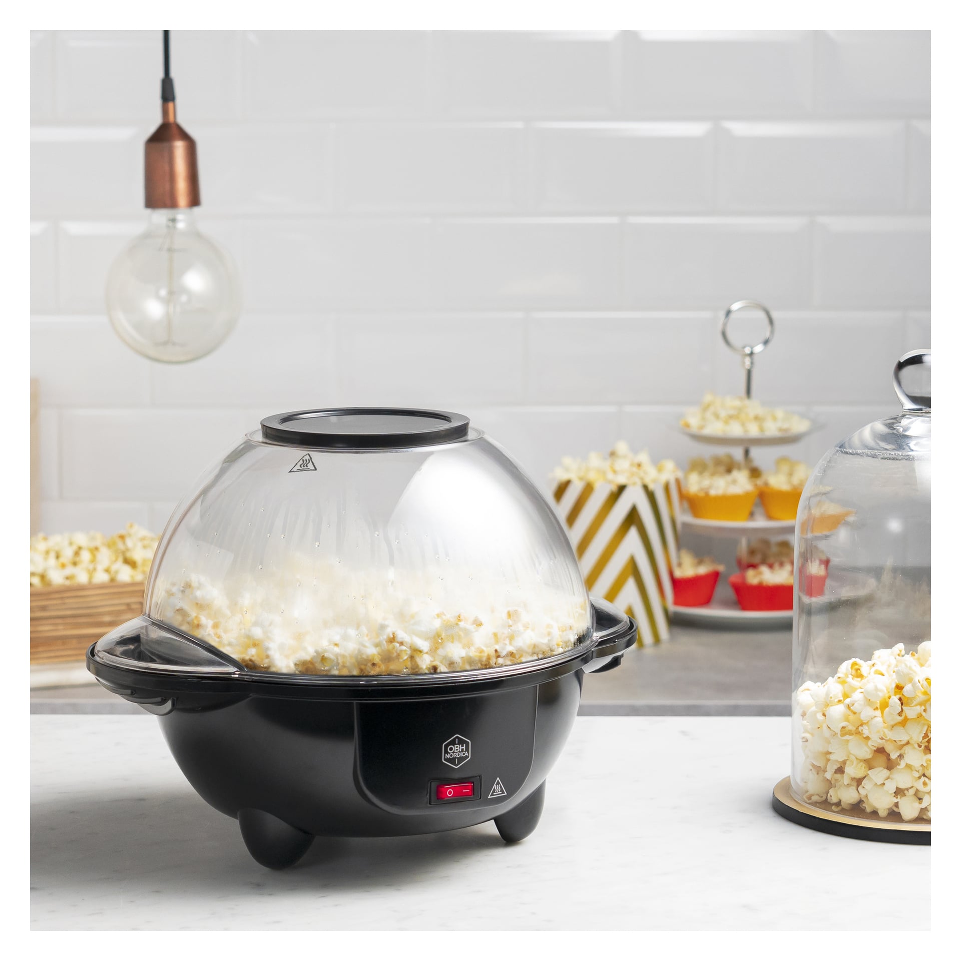 En svart popcornmaskin med ett genomskinligt kupollock står på köksbänken och poppar majs. Skålar och burkar med popcorn står bakom den.
