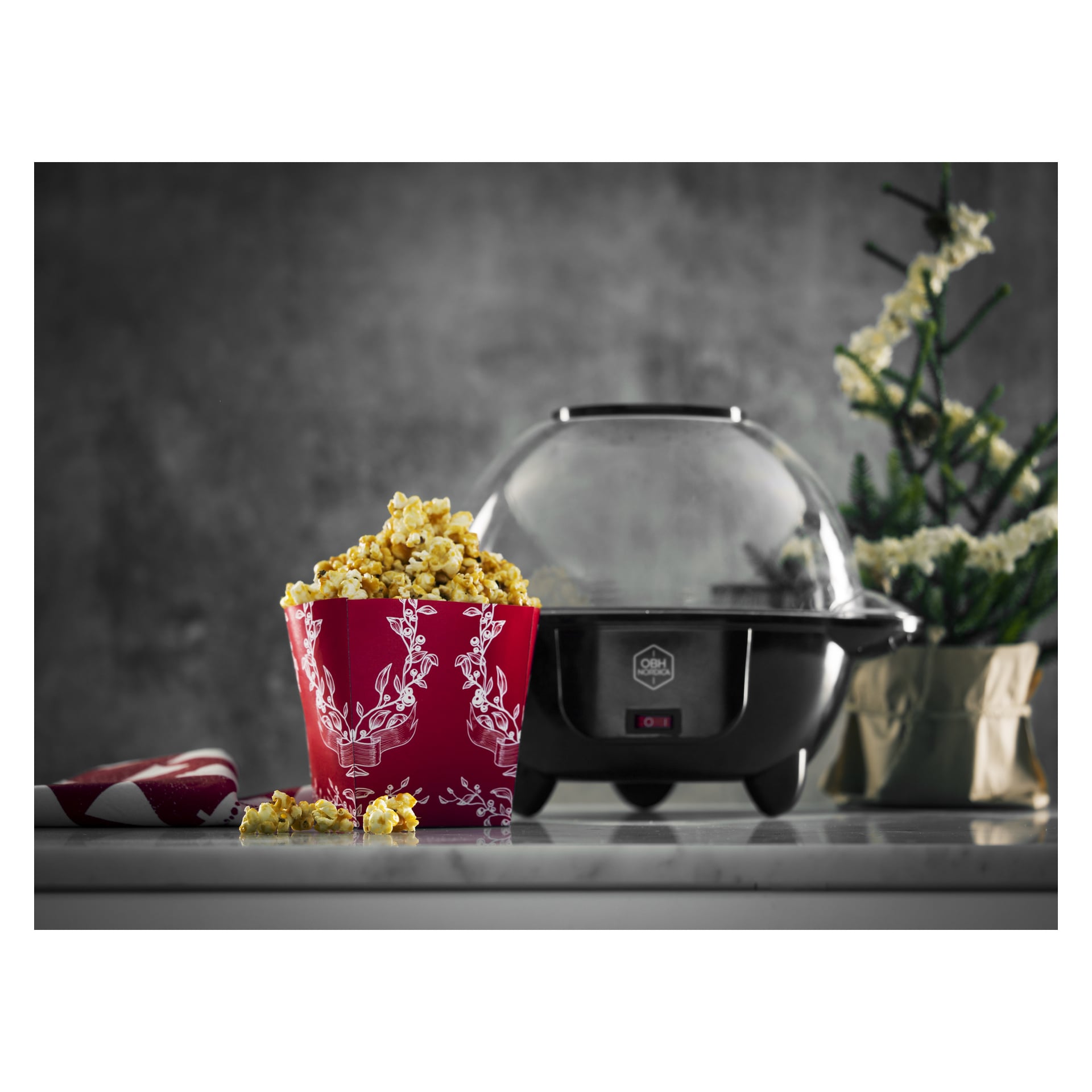 En svart popcornmaskin med genomskinligt lock står bredvid en röd popcornbehållare på en bänk, med en växt och en rödvit trasa bakom.