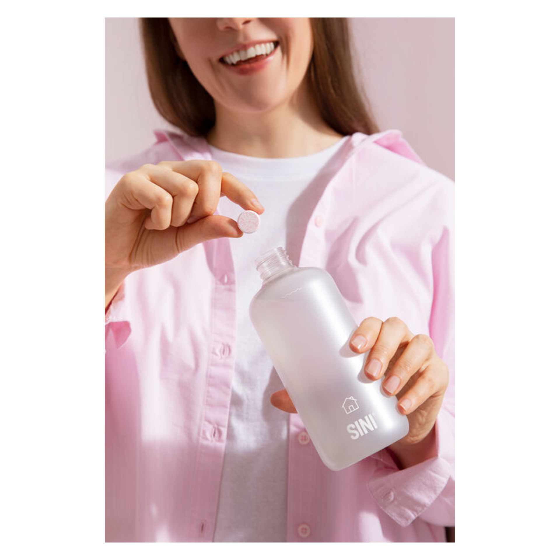 Någon i rosa skjorta och vit t-shirt håller en klar SINI-flaska och en rund tablett, redo att släppa ner tabletten i flaskan.