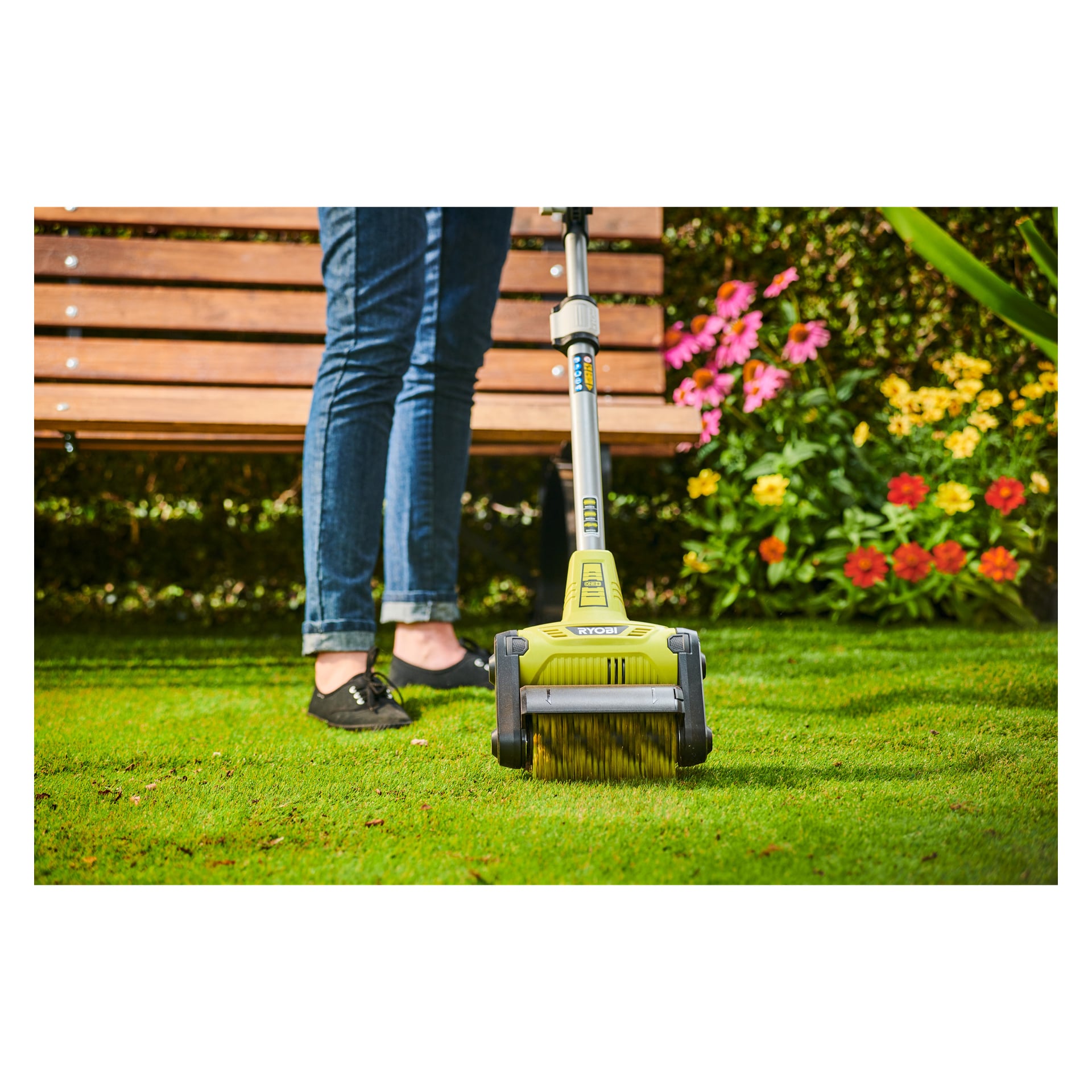 Någon i jeans och svarta skor använder en grön RAC823 rullborste på gräsmattan, med blommor, buskar och en träbänk bakom.
