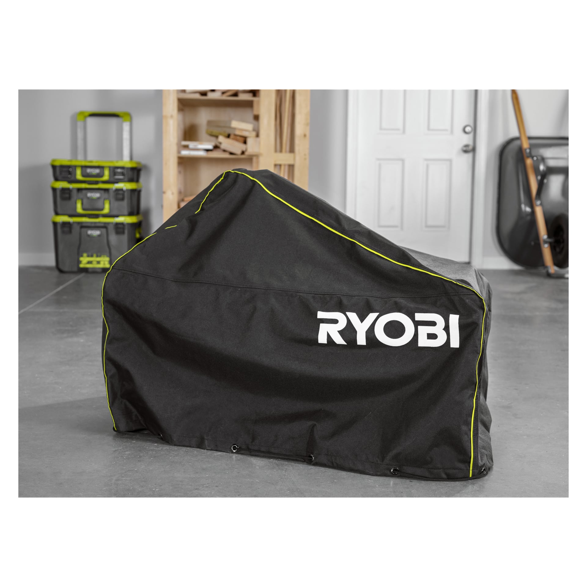 Ett svart RYOBI RAC658 snöslungeskydd skyddar utrustning i ett garage, omgiven av förvaringslådor och en närliggande vit dörr.