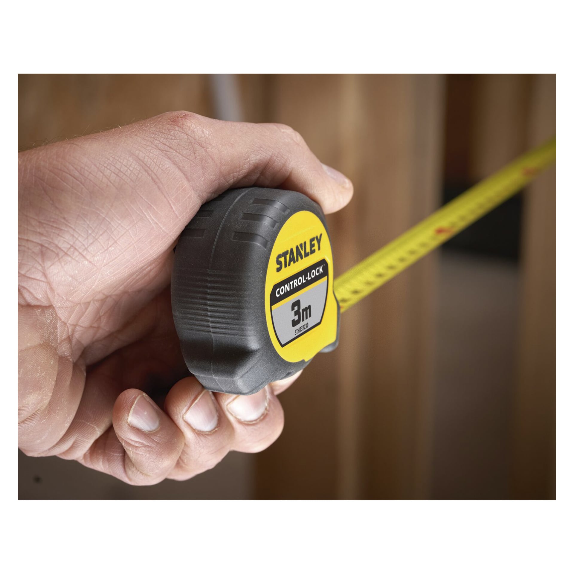 En hand håller ett Stanley Control-Lock-måttband som är utdraget till 3 meter, med suddiga träbalkar synliga i bakgrunden.