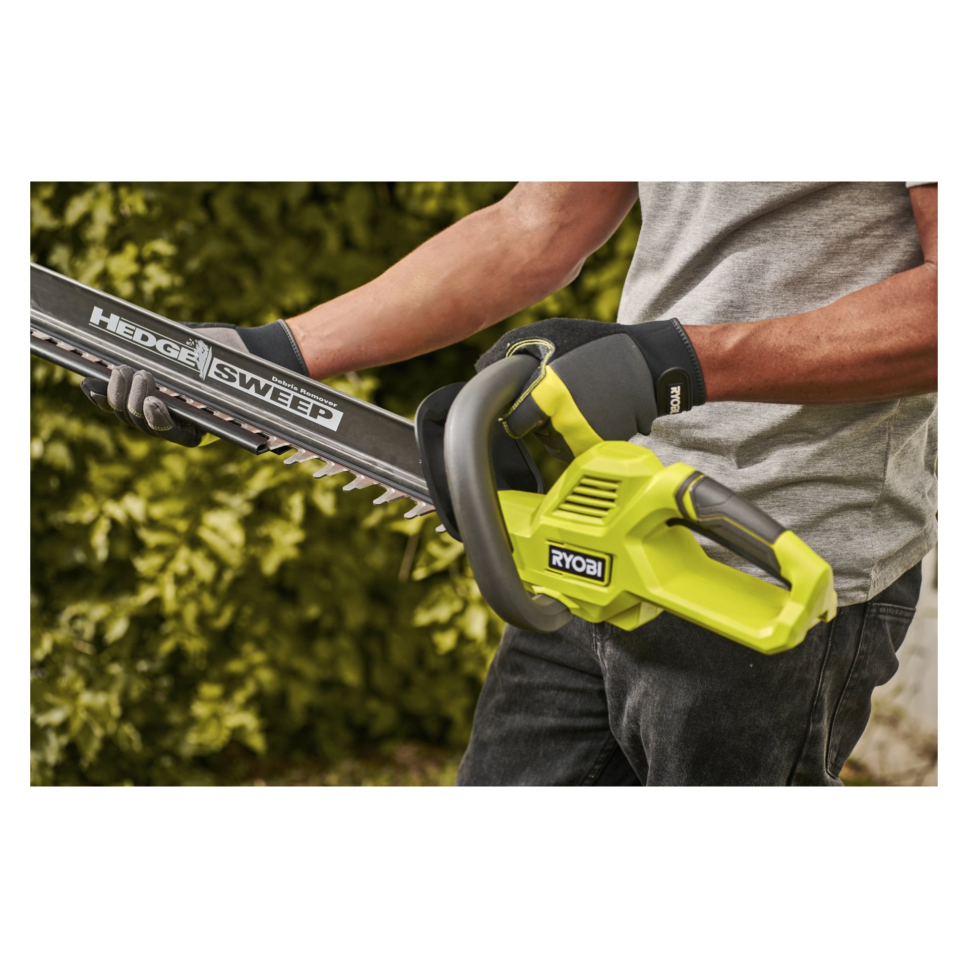 Handskbeklädda händer håller en gul Ryobi Hedge Sweep häcktrimmare utomhus, omgiven av frodigt grönt växtlighet i bakgrunden.