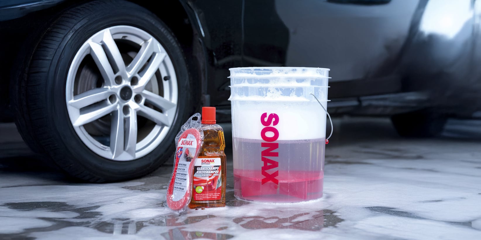 Sonax-autonpesushampoo, punainen kahvainen sieni ja saippuainen ämpäri ovat märällä marmorilla auton renkaan lähellä.