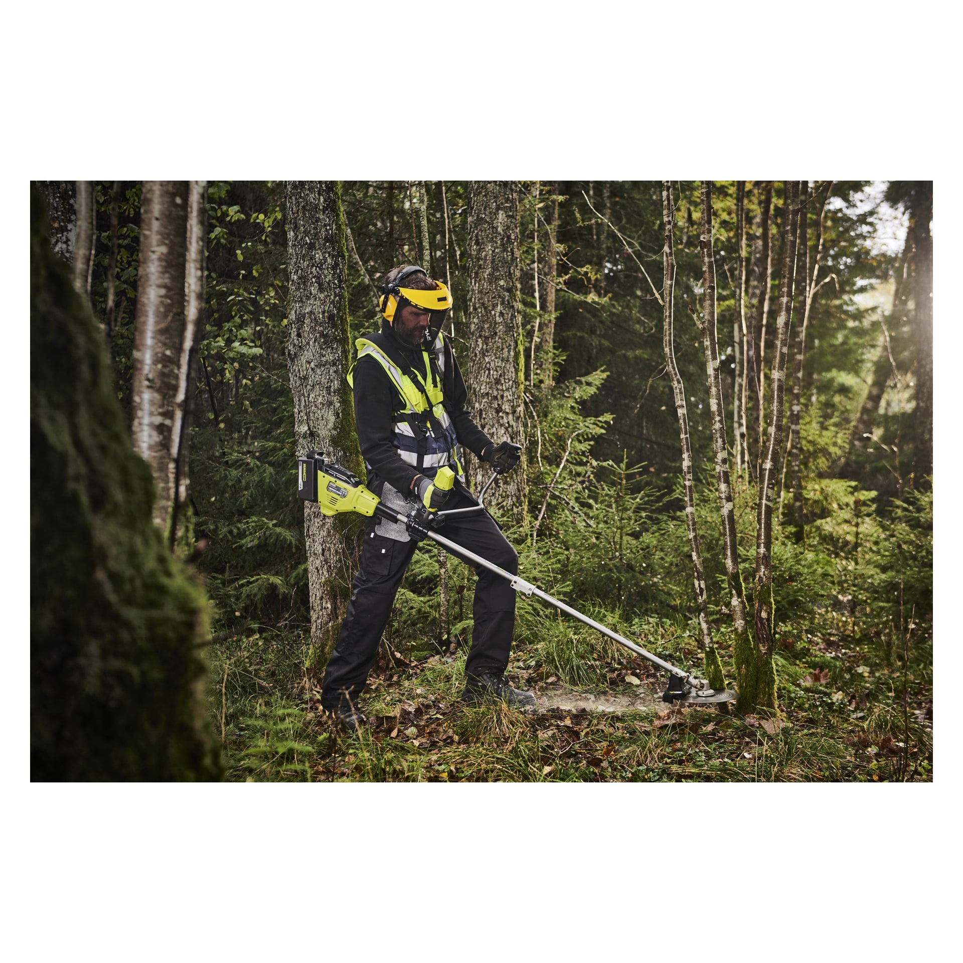 Med hjälm och reflektiv väst använder man en batteridriven buskklippare för att rensa skogsundervegetation i dagsljus.
