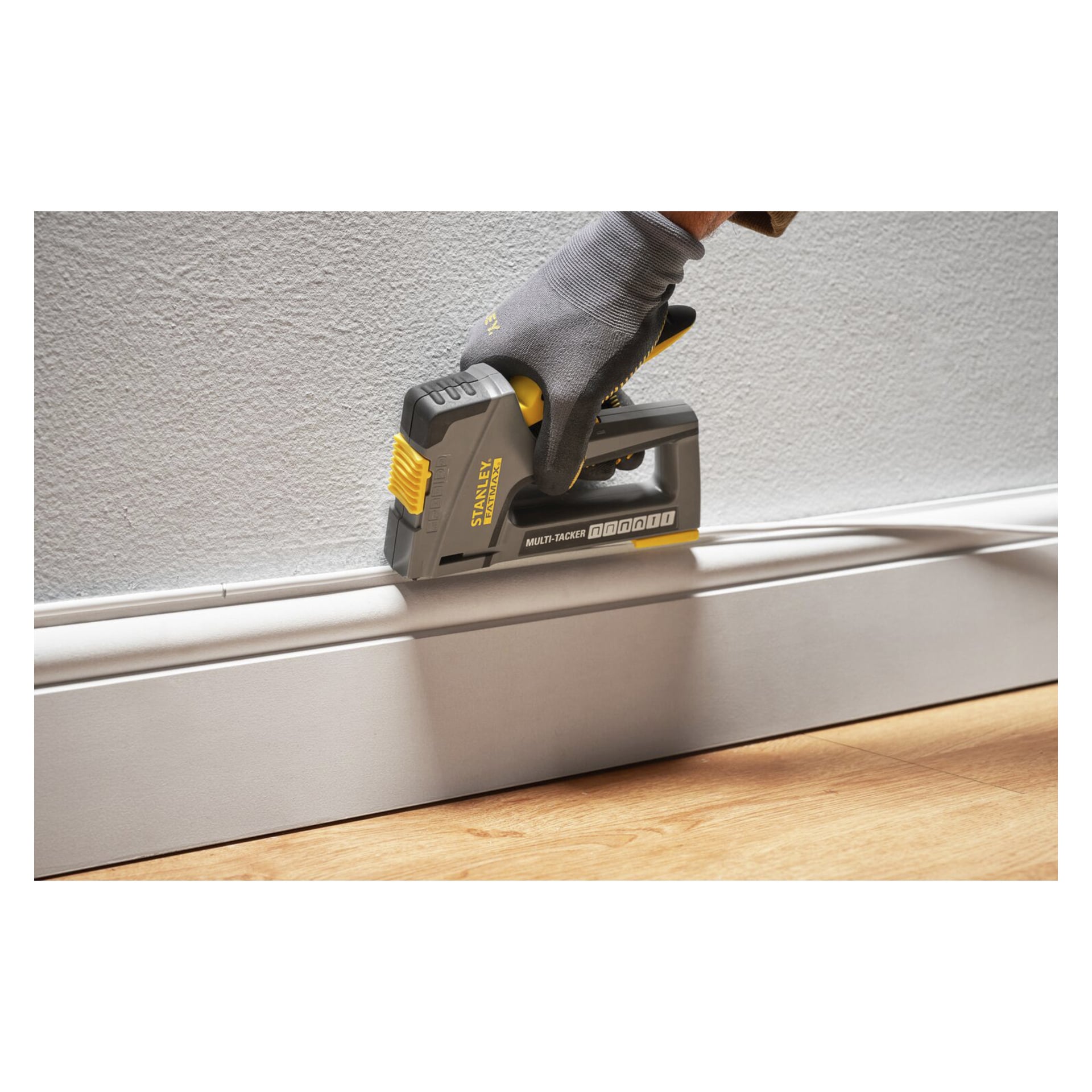 En handskbeklädd hand manövrerar ett Stanley FatMax Multi-Tacker-verktyg och fäster en kabel längs toppen av en vit golvliste.