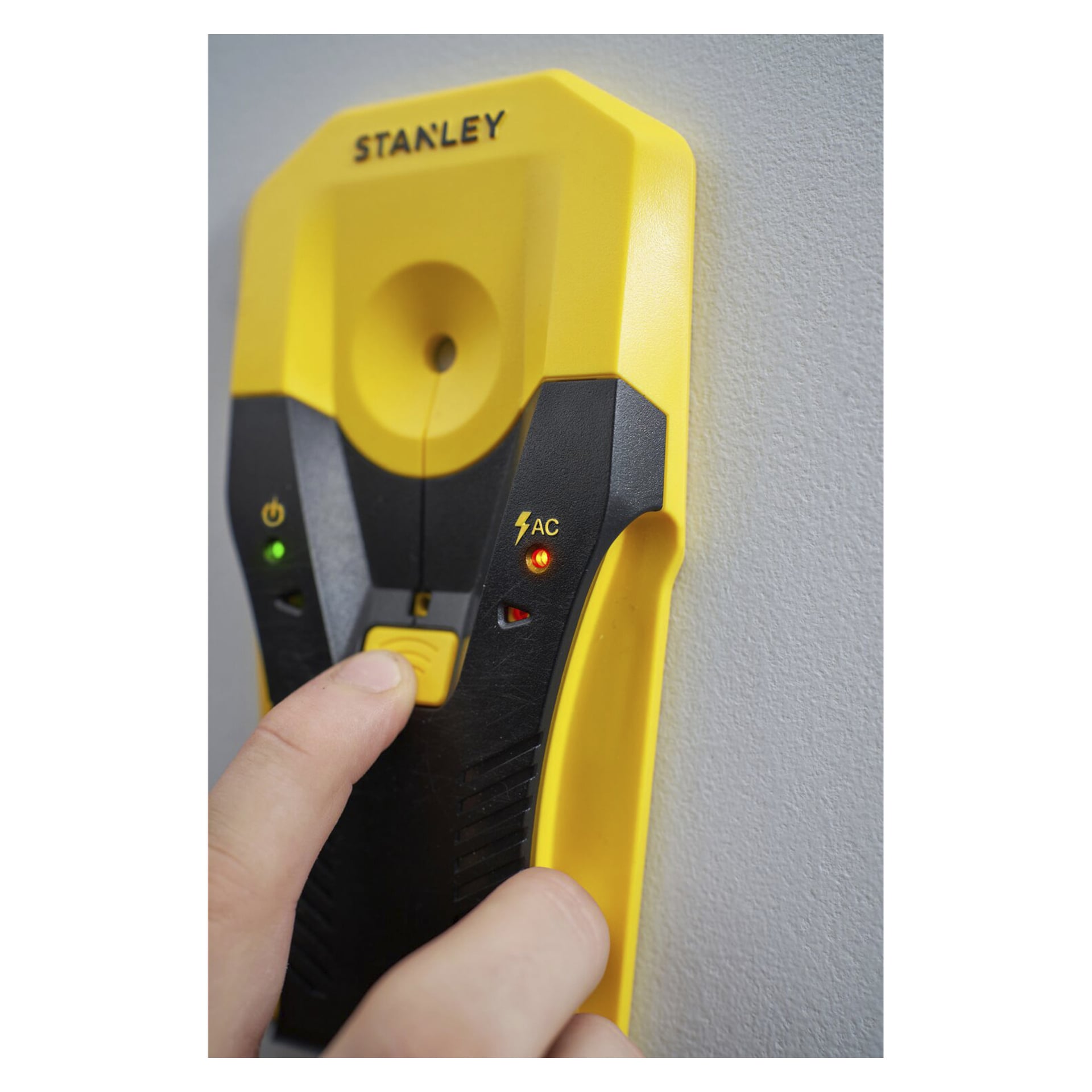En hand trycker på en gul knapp på en gul och svart Stanley regeldetektor monterad på en texturerad vägg. AC-indikatorlampan är röd.
