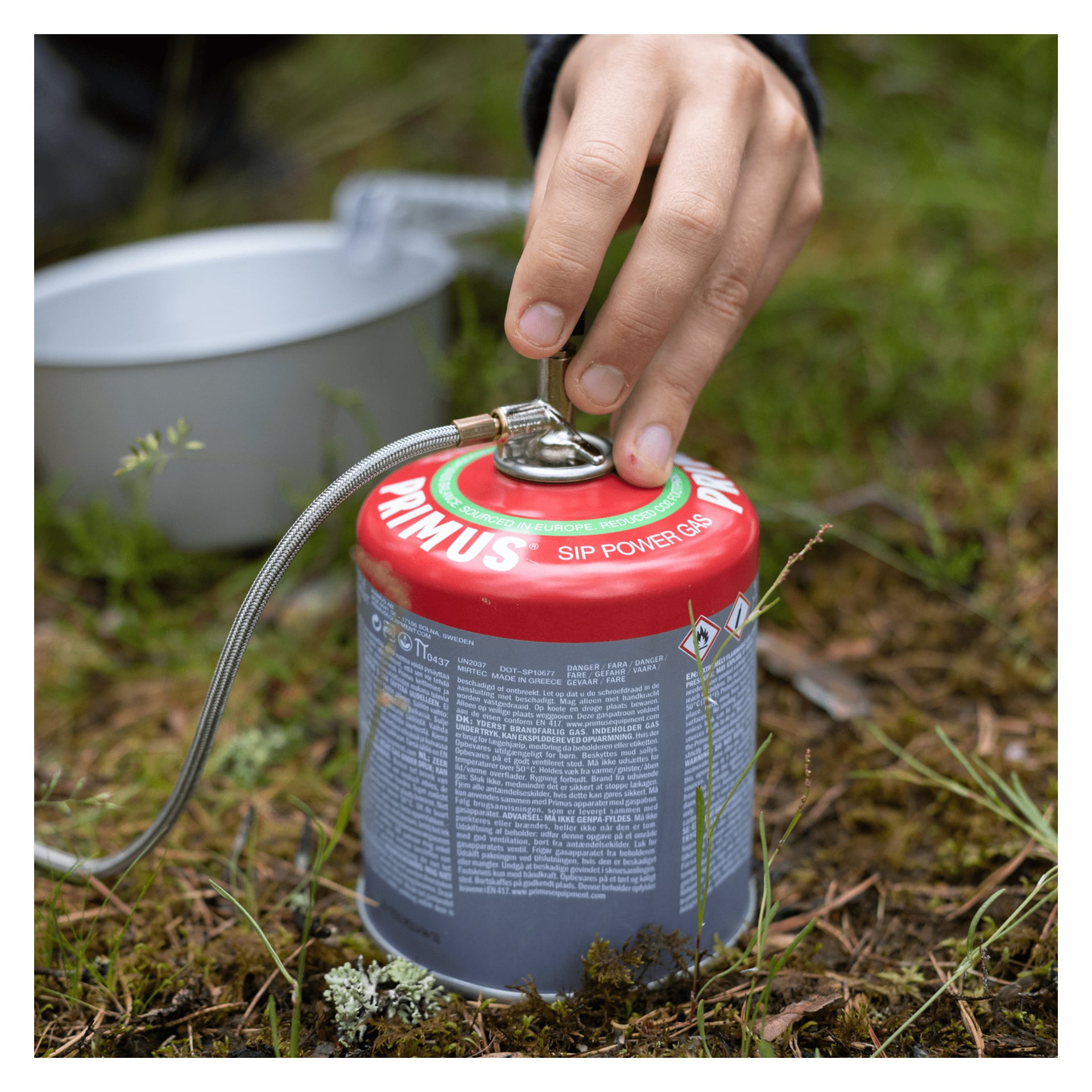 En hand vrider ventilen på en röd och grå Primus-gasbehållare med bränsleslang, som vilar på gräset bredvid en metallburk utomhus.